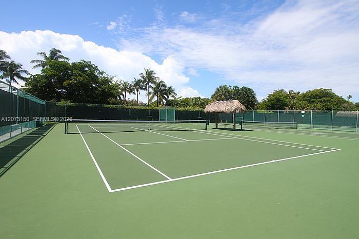 Private tennis court featuring a hard surface, perimeter chain-link fencing with wind-screen paneling, and a thatched-roof palapa structure