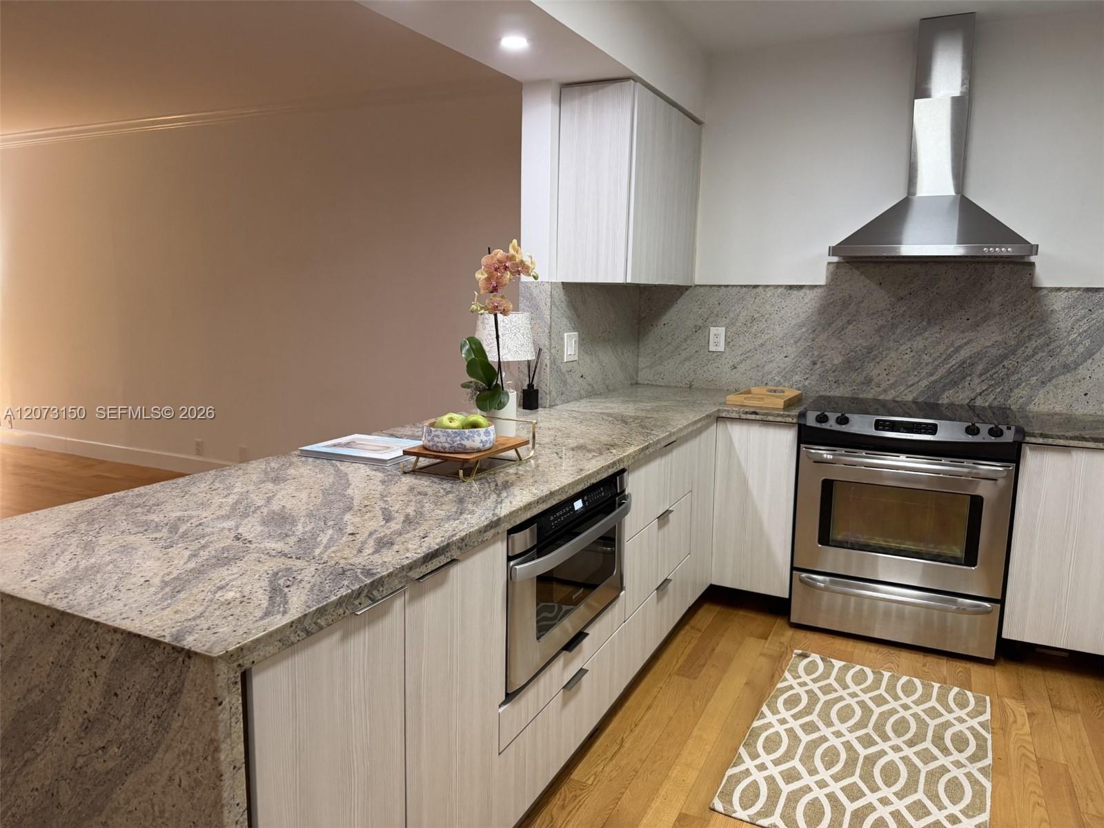 Kitchen featuring flat-panel cabinetry, stone countertops, stainless steel appliance suite, chimney-style range hood, and wood-finish flooring