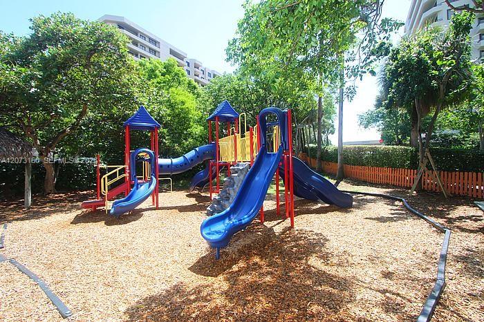 Outdoor play area featuring multi-level slides, a tunnel attachment, climbing structure, wood chip ground cover, and mature shade trees