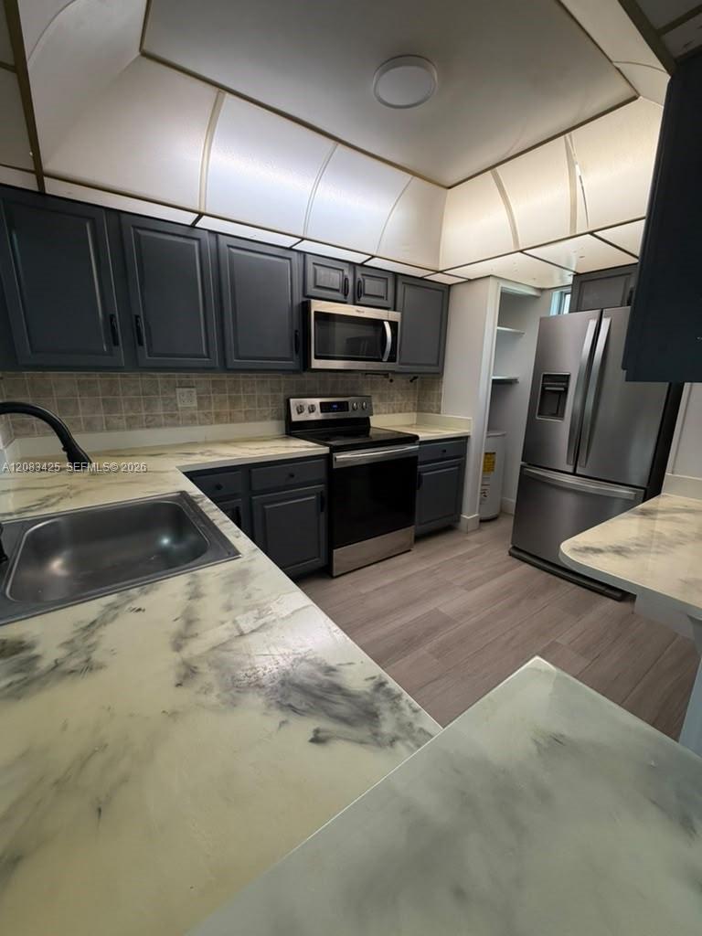 Kitchen featuring dark-toned cabinetry, stainless steel appliances, patterned countertops, wood-finish flooring, and a tray ceiling with recessed lighting Kitchen featuring dark-toned cabinetry, stainless steel appliances, patterned countertops, wood-finish flooring, and a tray ceiling with recessed lighting
