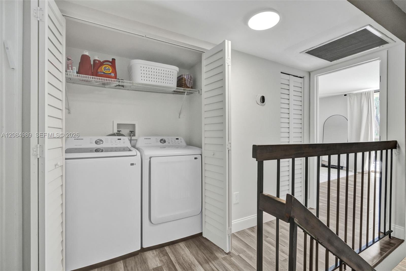 Laundry area featuring louvered bi-fold closet doors, integrated wire shelving, and wood-finish flooring with a metal stair railing