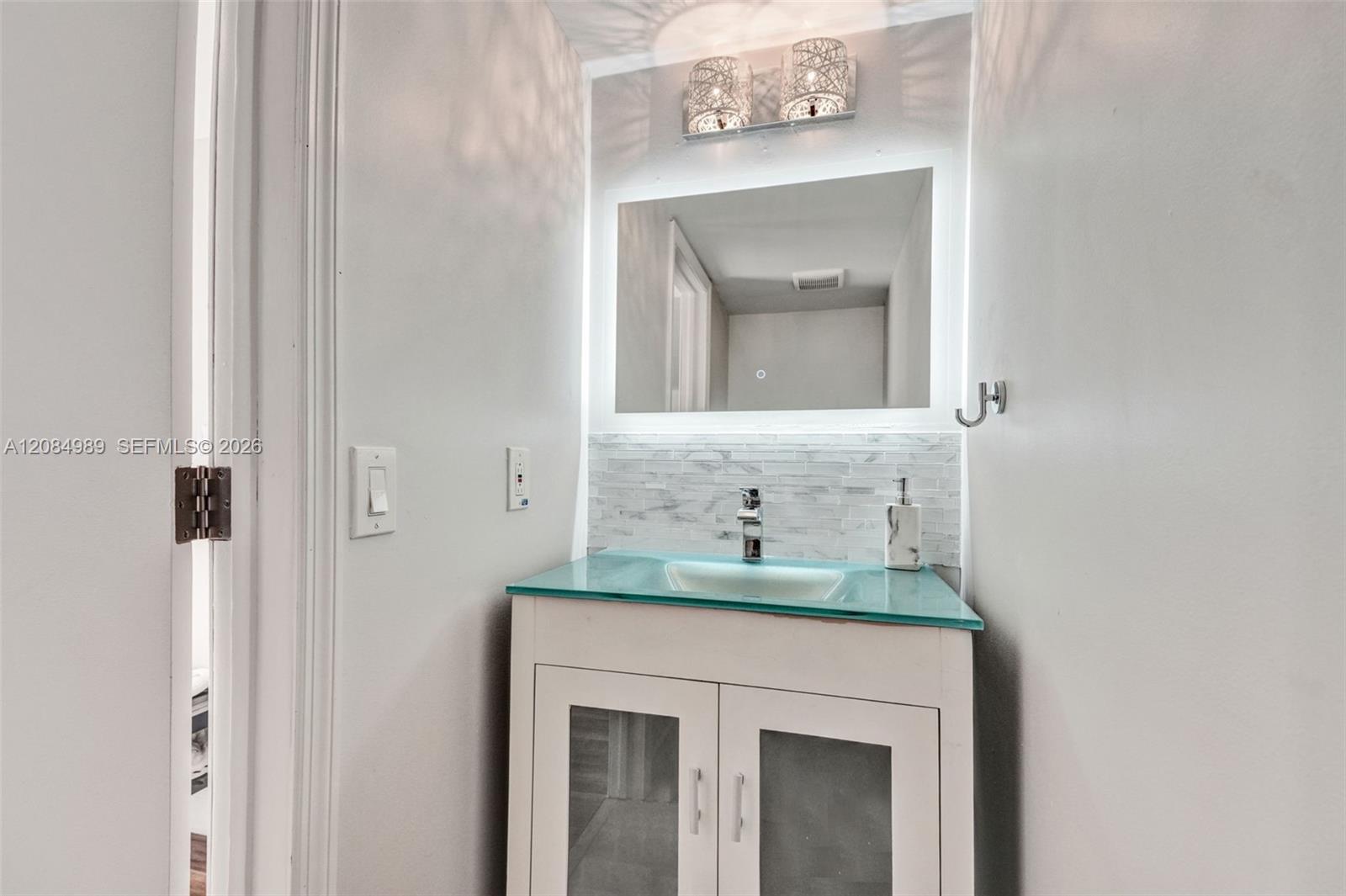Vanity featuring a glass countertop with an integrated basin, white cabinetry with frosted glass inserts, LED-backlit mirror, and a textured tile backsplash