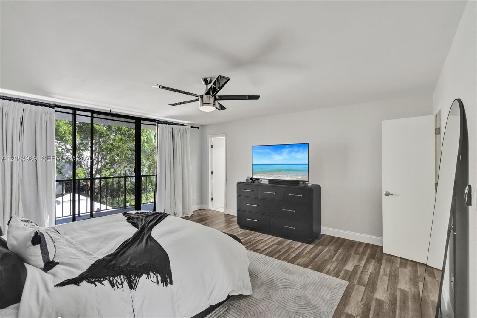 Floor-to-ceiling glass sliding doors with black framing, wood-finish flooring, metal ceiling fan with integrated lighting, and access to a railed balcony