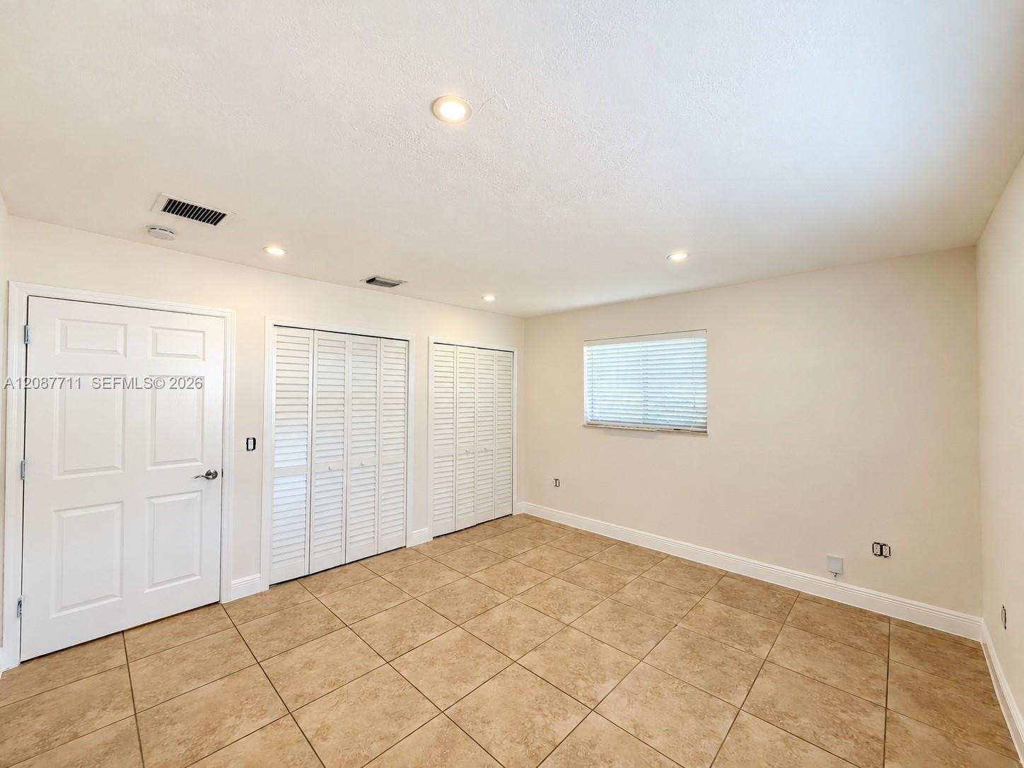 Room featuring neutral tile flooring, recessed ceiling lighting, two sets of white louvered-style closet doors, and a single window with integrated blinds