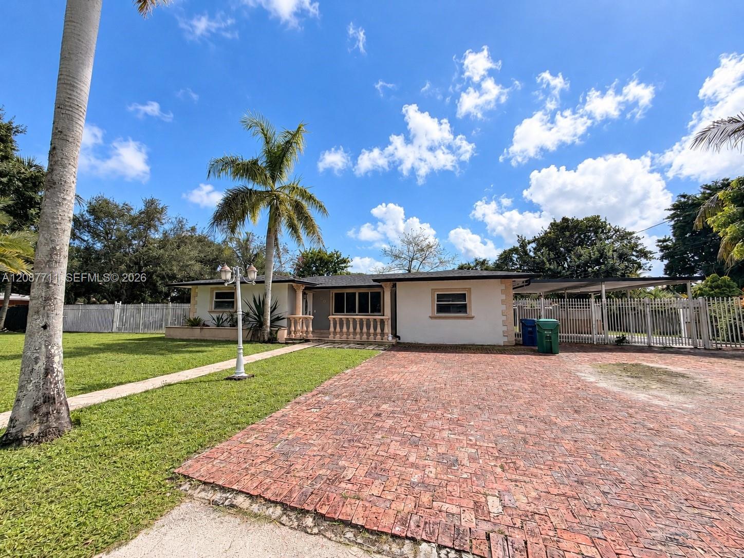 Single-story exterior featuring a brick-paved driveway, decorative balustrade porch railing, attached carport, and perimeter metal fencing