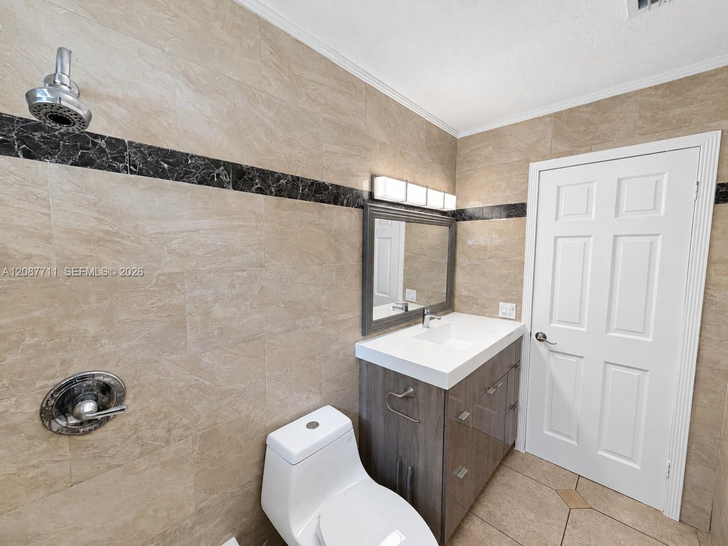 Full-wall tile surround with dark stone accent banding, wood-finish vanity with integrated sink, four-light vanity fixture, and six-panel interior door