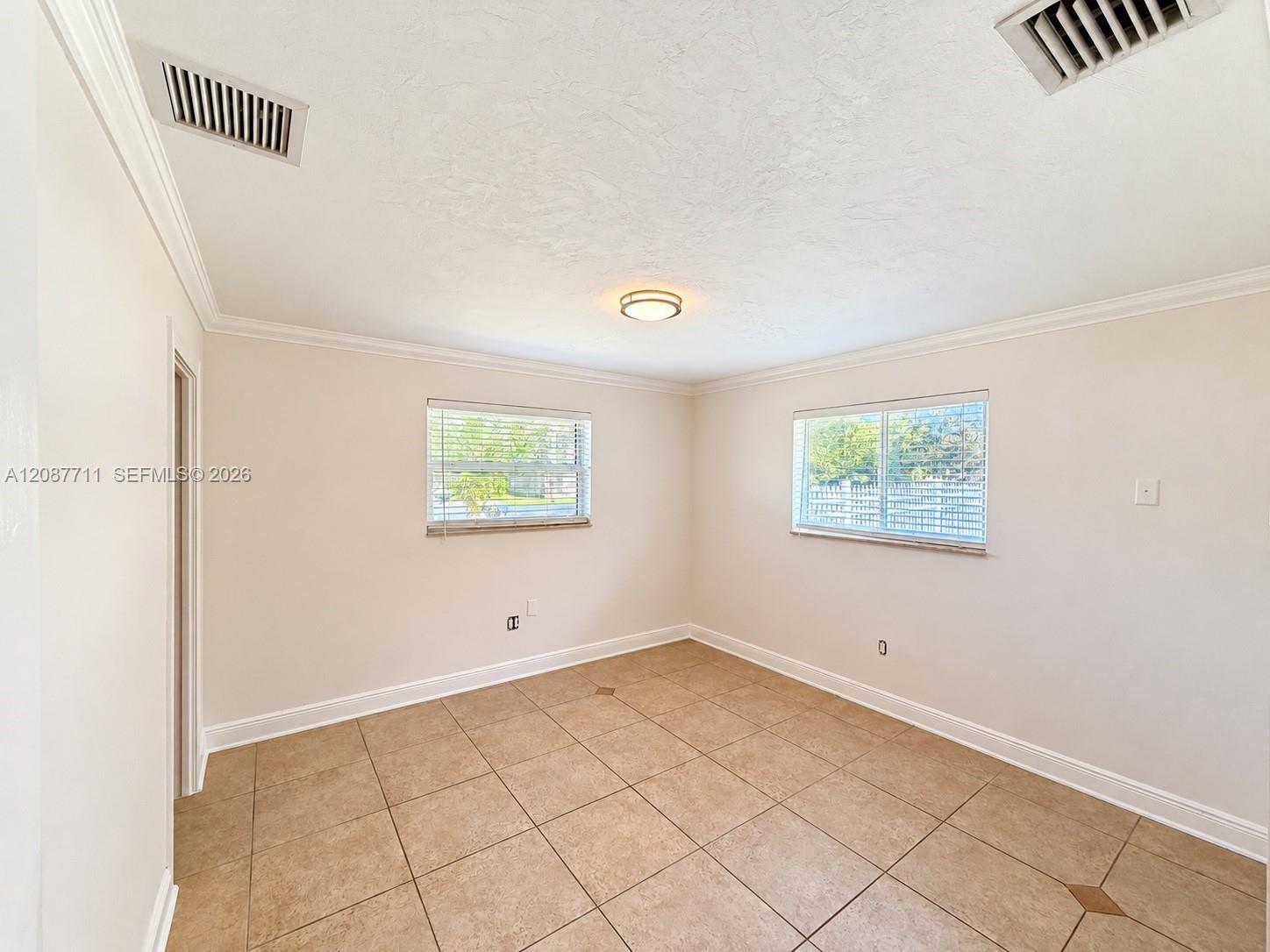 Neutral-toned interior featuring large-format square tile flooring, crown molding, dual windows with horizontal blinds, and a centered flush-mount ceiling fixture