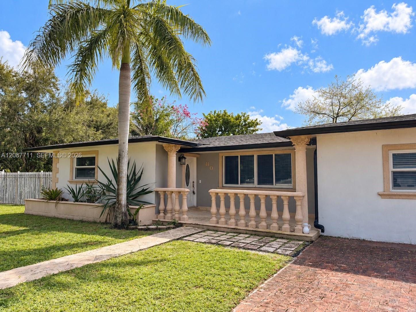 Stucco exterior featuring a covered front porch with decorative balustrade and columns, brick-paved driveway, and mature landscaping