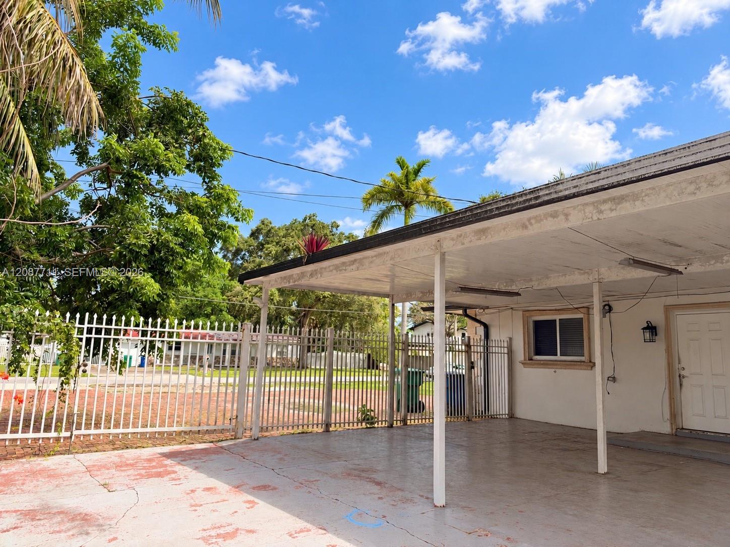 Covered carport with concrete flooring, exterior wall-mounted light fixture, and perimeter metal fencing