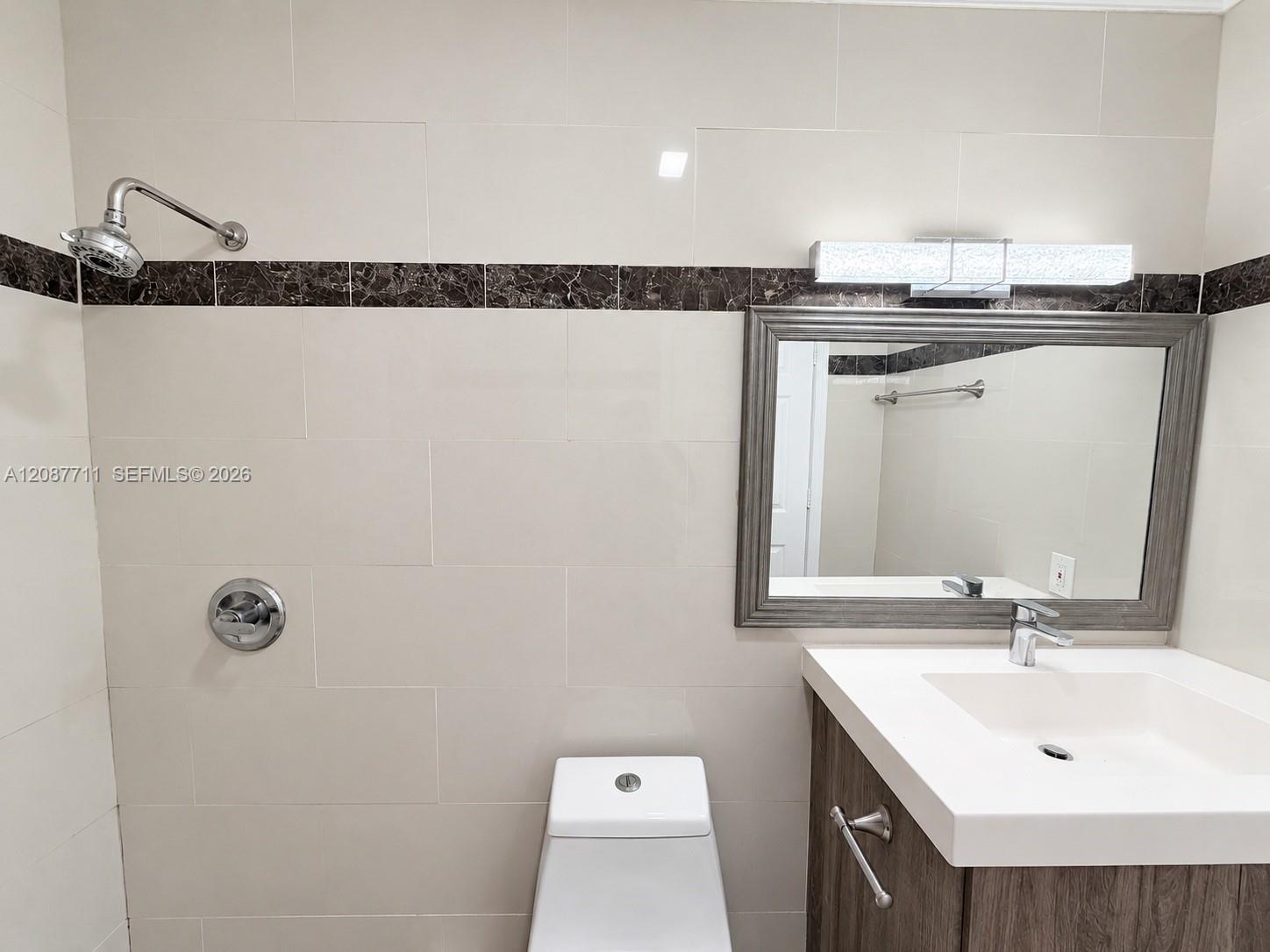 Bathroom featuring floor-to-ceiling cream tiling with dark stone accent banding, a vanity with an integrated basin, wall-mounted brushed-metal hardware, and a linear light fixture