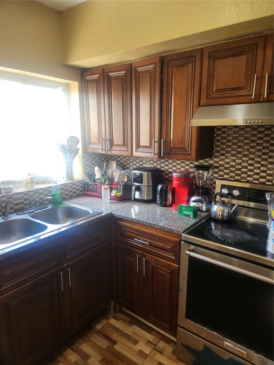 Kitchen featuring stained wood cabinetry, stone-finish countertops, stainless steel dual-basin sink, patterned tile backsplash, and stainless steel range Kitchen featuring stained wood cabinetry, stone-finish countertops, stainless steel dual-basin sink, patterned tile backsplash, and stainless steel range