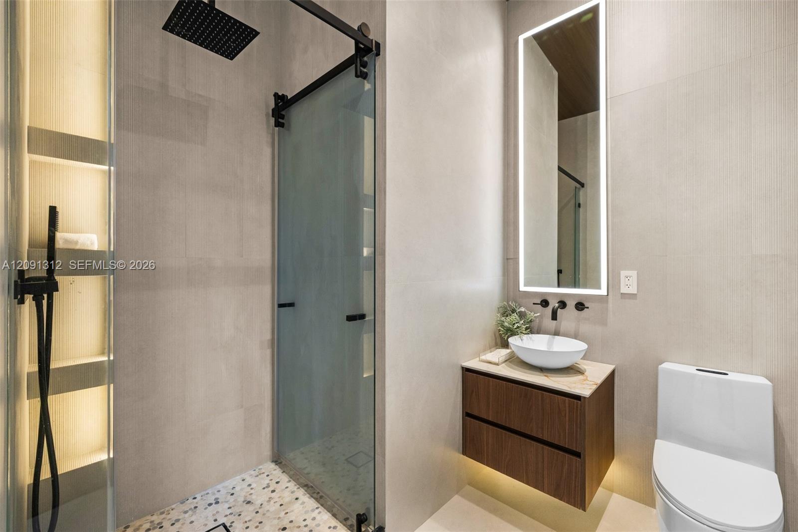 Floating wood-finish vanity with a stone countertop and white vessel sink, large-format tile wall cladding, walk-in shower with matte black hardware and sliding glass door, pebble-stone shower floor, and back-lit rectangular mirror