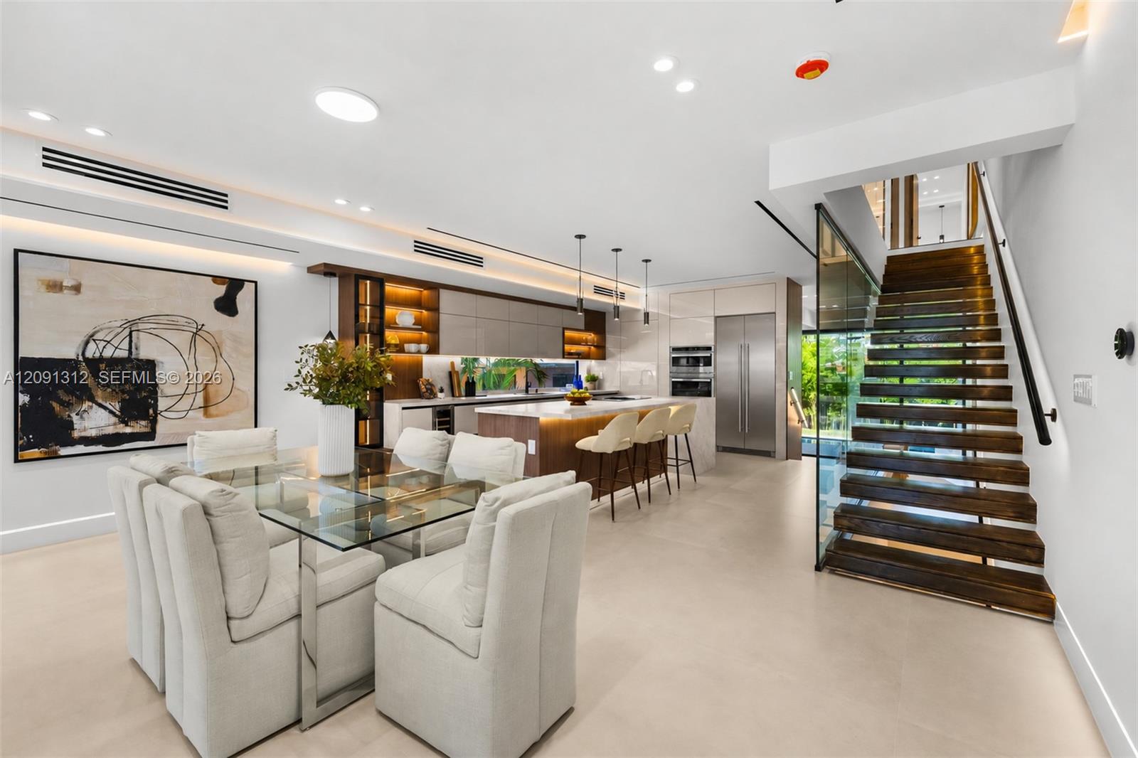 Modern kitchen featuring a stone-topped island with integrated cabinetry, built-in stainless steel appliances, recessed linear lighting, and a floating wood staircase with glass paneling