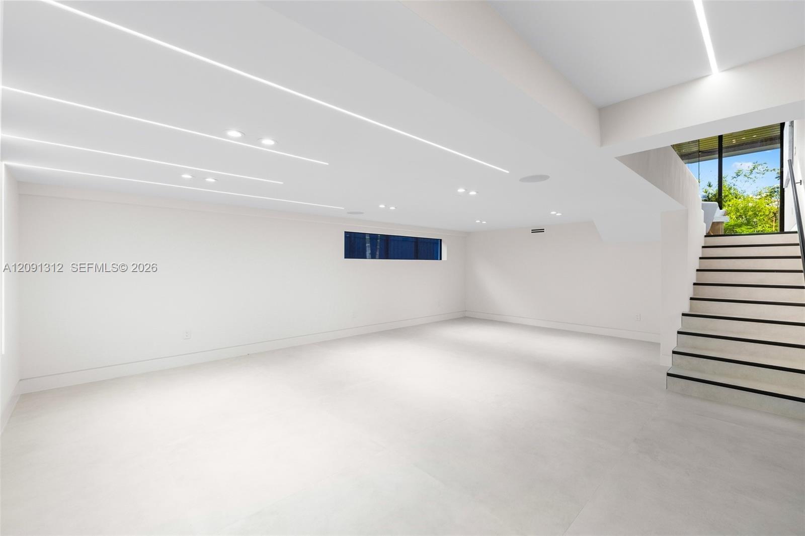 Open floor plan featuring recessed linear lighting, smooth white walls, large-format tile flooring, and a staircase with a metal handrail