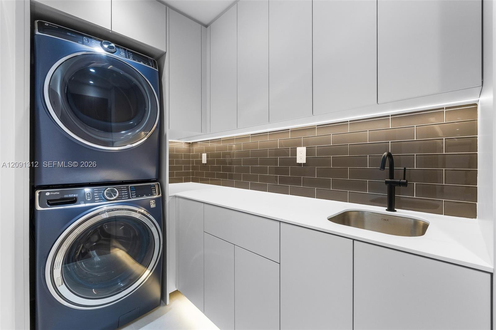 Laundry area featuring stacked front-load appliances, quartz countertops, high-gloss cabinetry, a stainless steel undermount sink with a matte black faucet, and a tiled backsplash