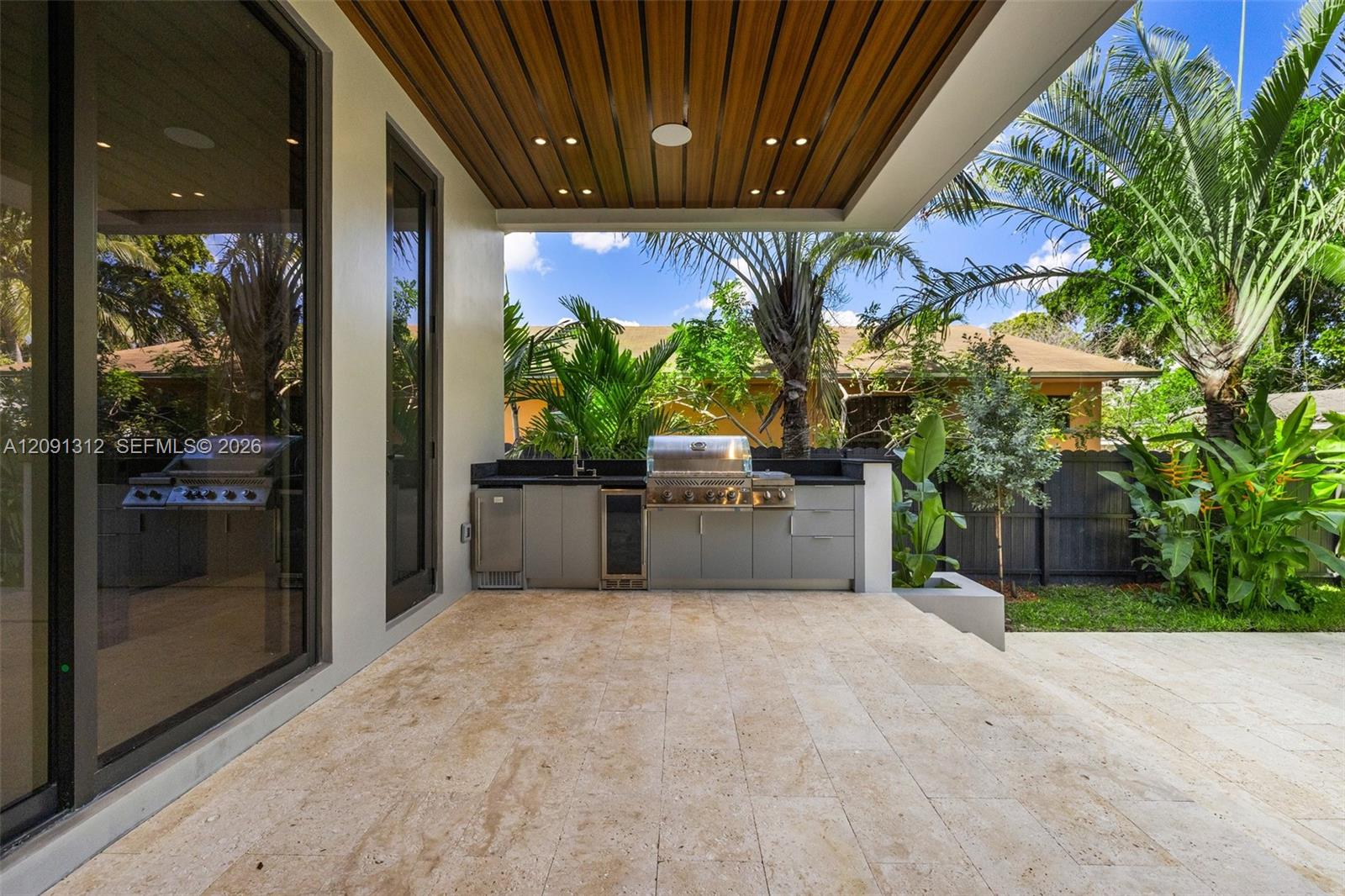 Covered outdoor kitchen featuring built-in cabinetry, stainless steel grill, beverage cooler, stone-finish flooring, and a wood-slat ceiling