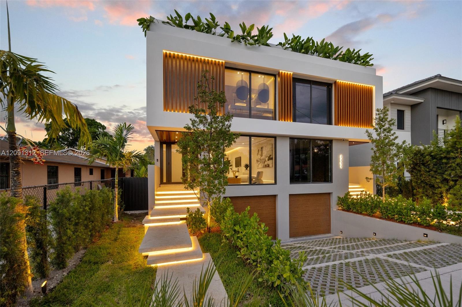 Modern two-story architectural exterior featuring smooth white stucco finish, vertical wood-slat accents, integrated LED stair and facade lighting, large-format glass sliders, and a permeable paver driveway