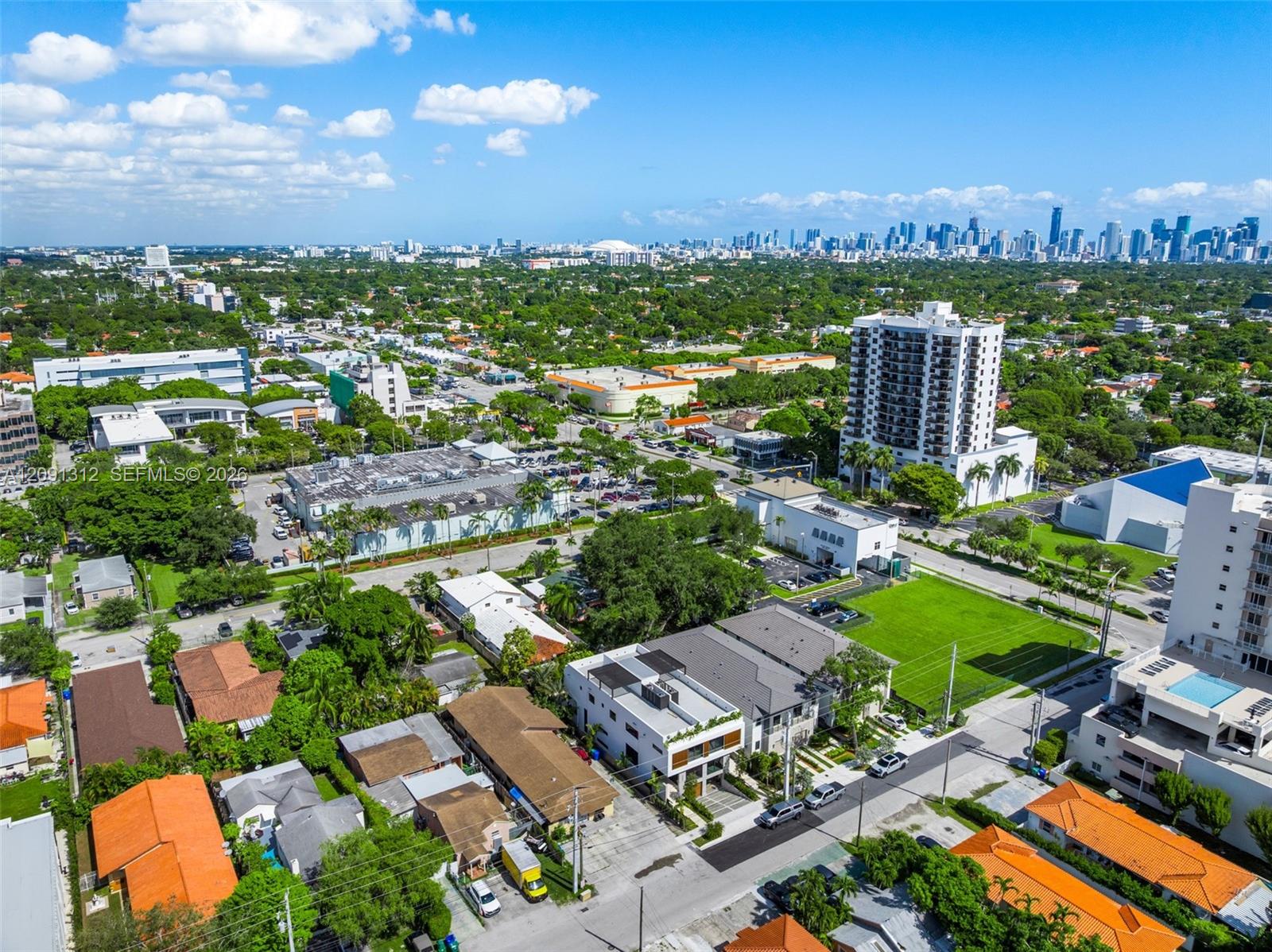 Aerial perspective of urban landscape featuring high-rise city skyline, mature tree canopy, multi-family residential structures, and paved street infrastructure