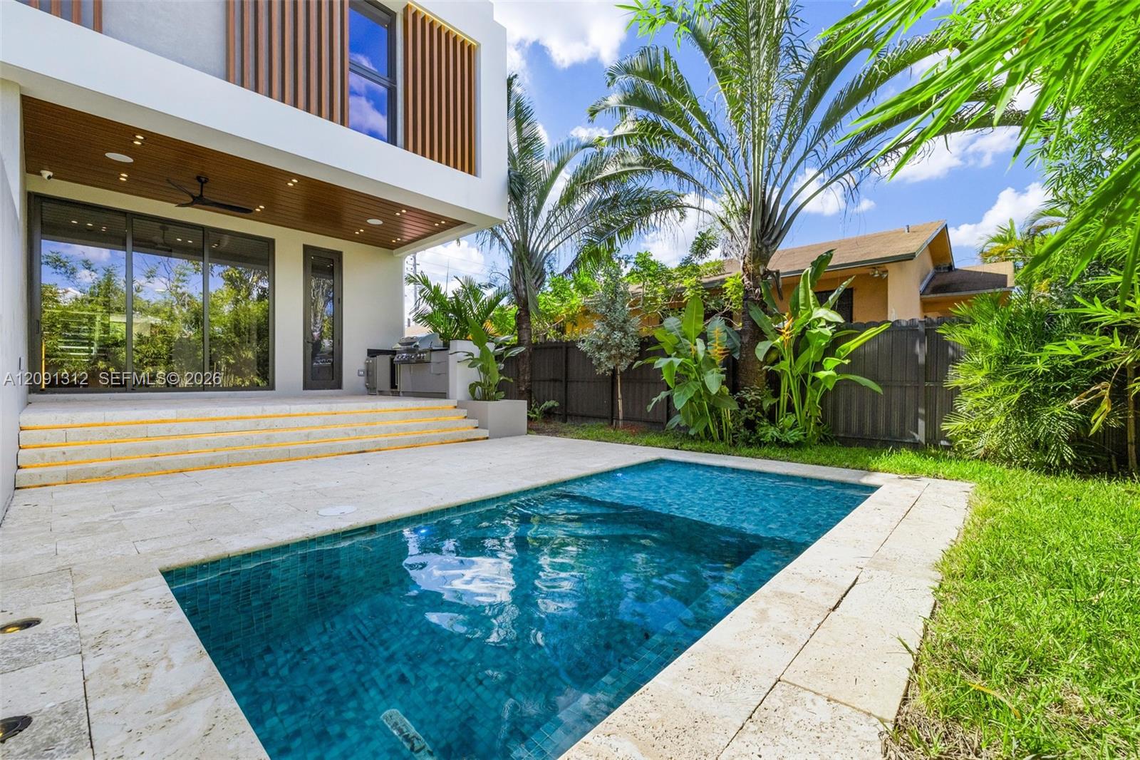 Modern exterior featuring a swimming pool, travertine deck, built-in outdoor kitchen, wood-panel soffit, and floor-to-ceiling sliding glass doors