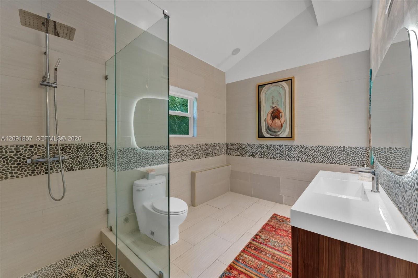 Bathroom featuring a glass-enclosed shower with pebble stone flooring and wall accents, a wood-finish vanity with a rectangular sink, wall-mounted illuminated mirrors, and a vaulted ceiling