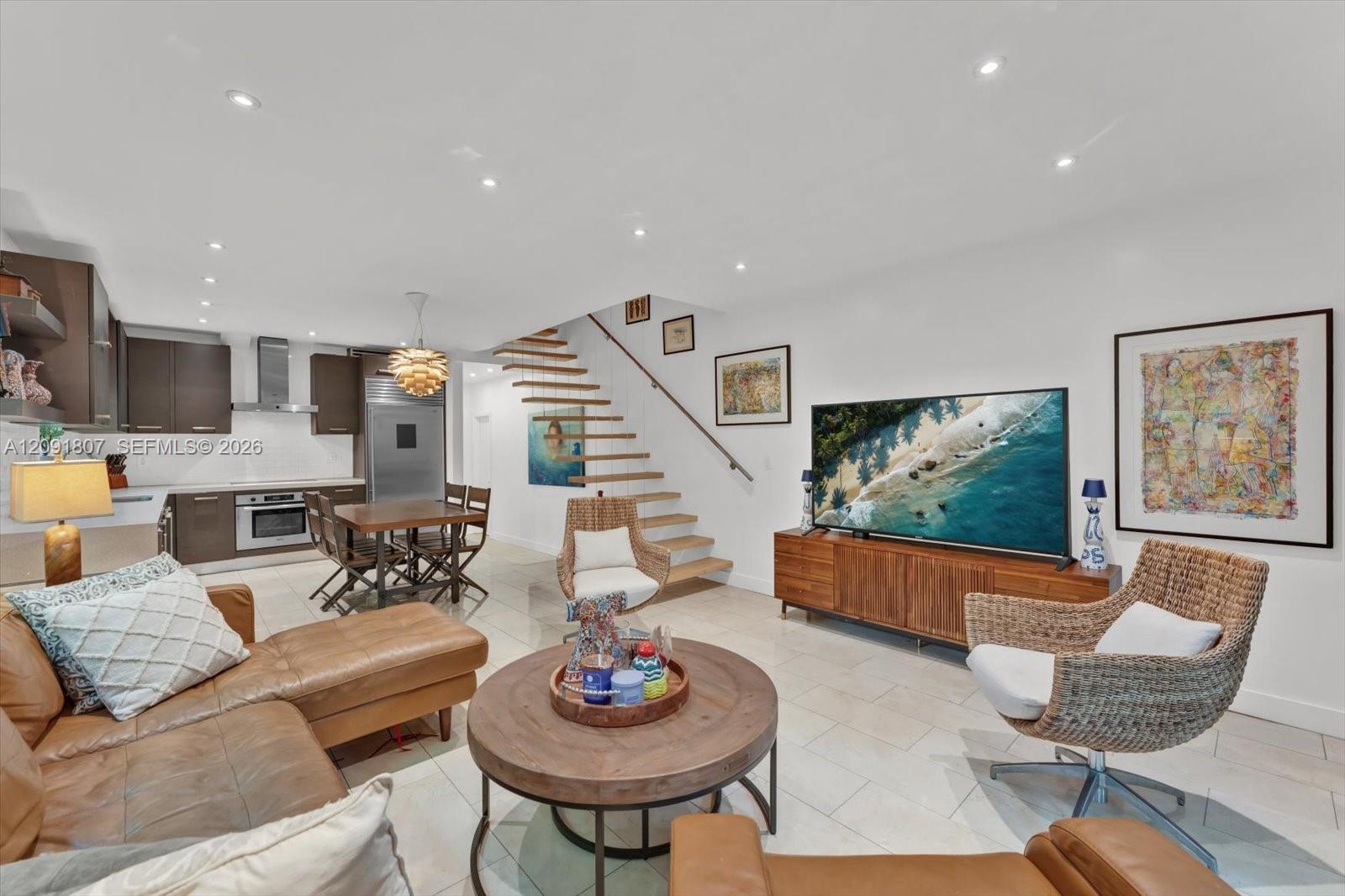 Open-concept area featuring recessed ceiling lighting, light-toned tile flooring, custom cabinetry, and a floating staircase with a metal handrail