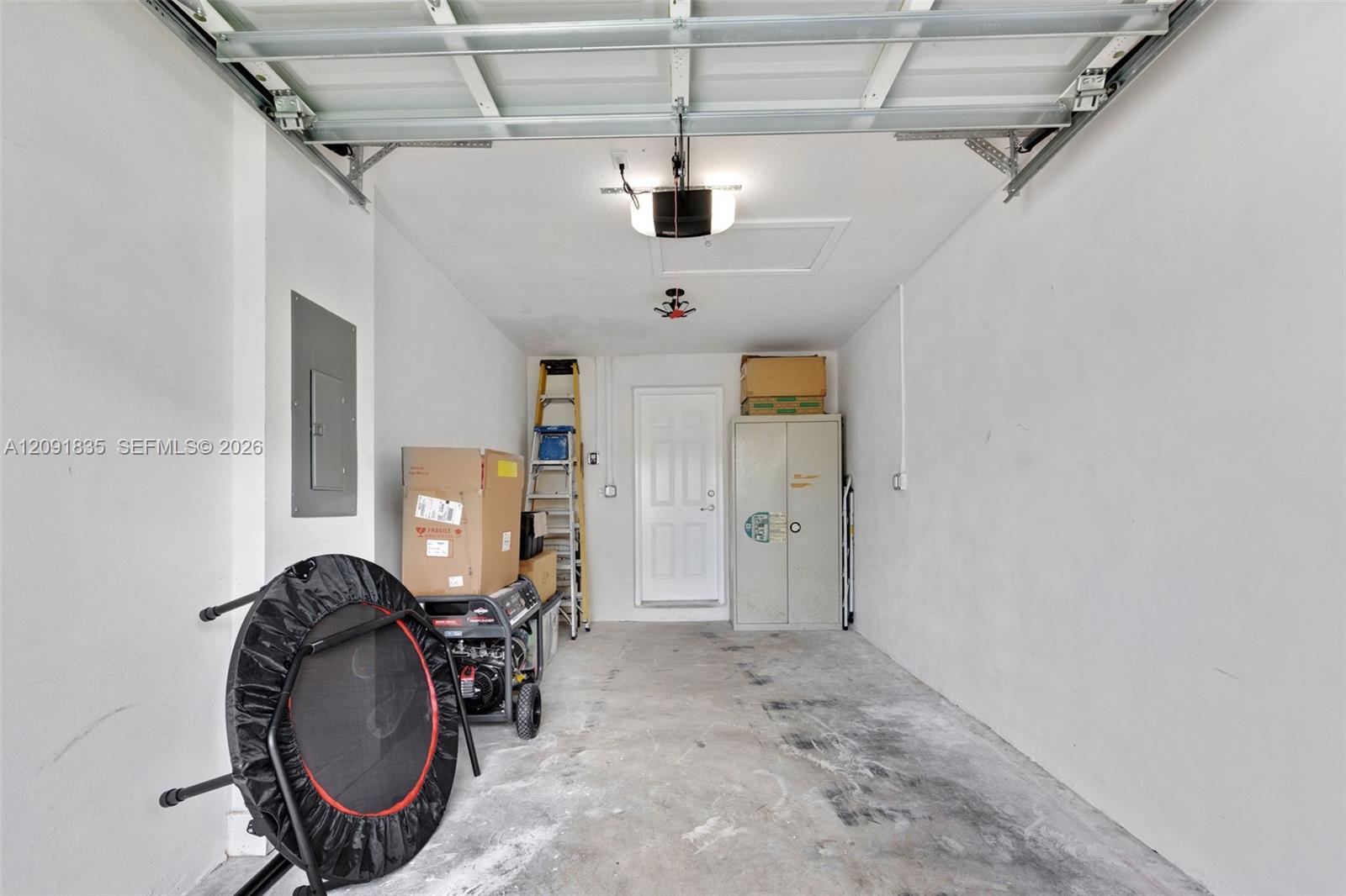 Garage interior featuring a concrete floor, automated overhead door system, ceiling-mounted lighting, integrated electrical panel, and a paneled service door