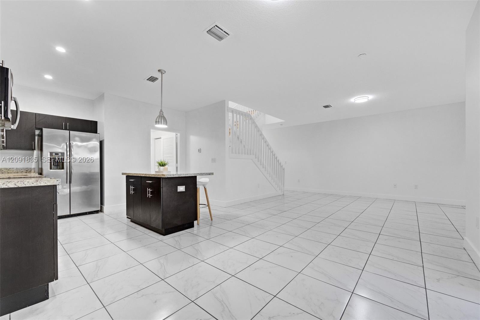 Open-concept area featuring white tile flooring, dark wood-finish cabinetry, stone countertops, a center island with pendant lighting, and a white staircase with wooden balusters