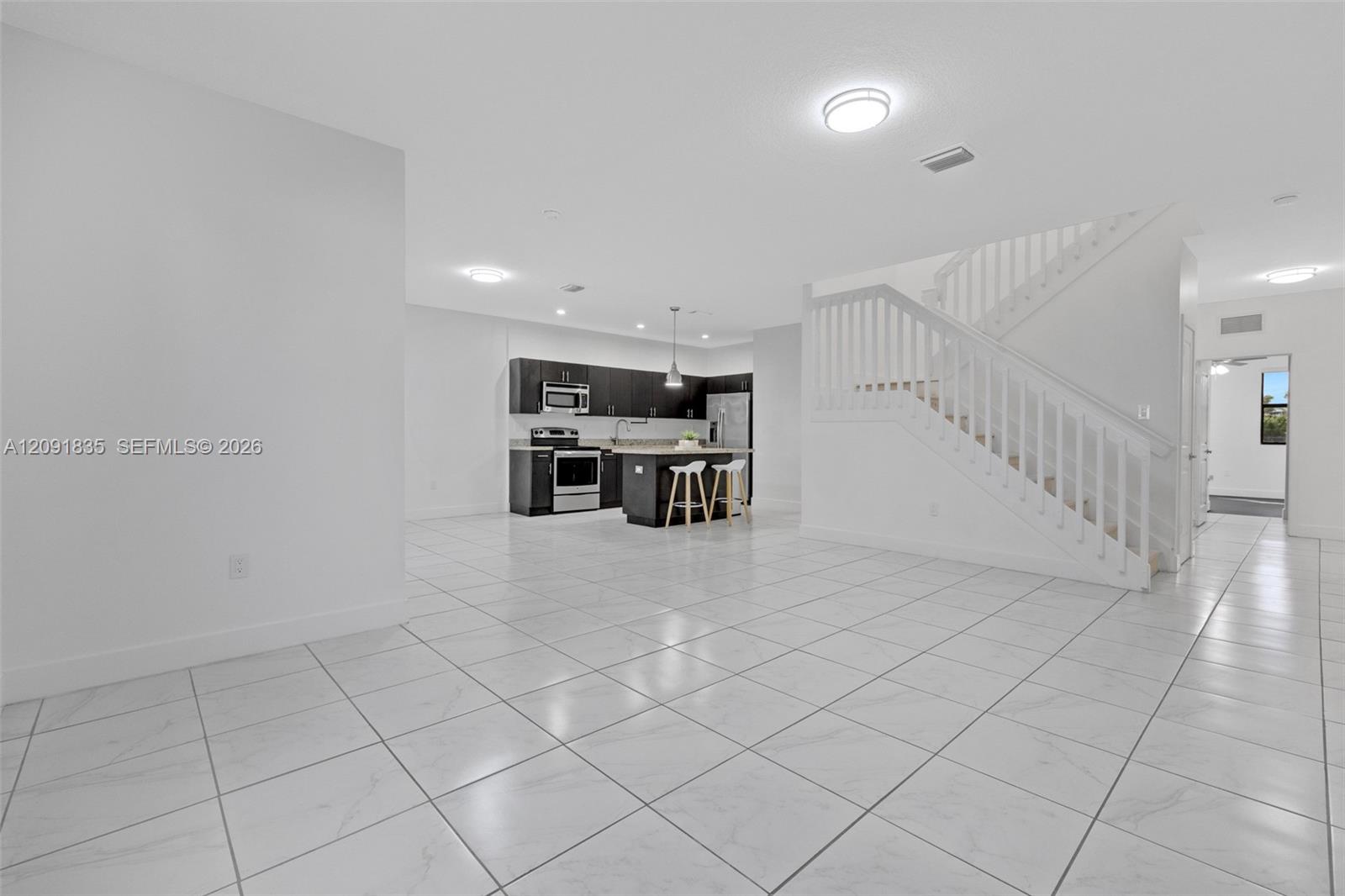 Open-concept interior featuring light-toned tile flooring, kitchen with dark cabinetry, central island, and staircase with white railings