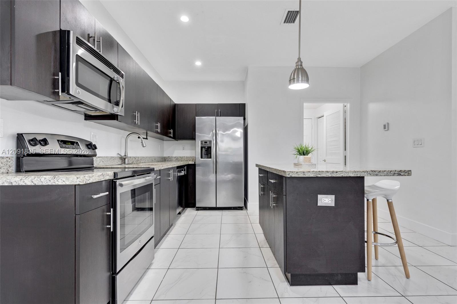 Kitchen featuring flat-panel dark cabinetry, speckled stone-finish countertops, stainless steel appliances, a center island with pendant lighting, and ceramic tile flooring