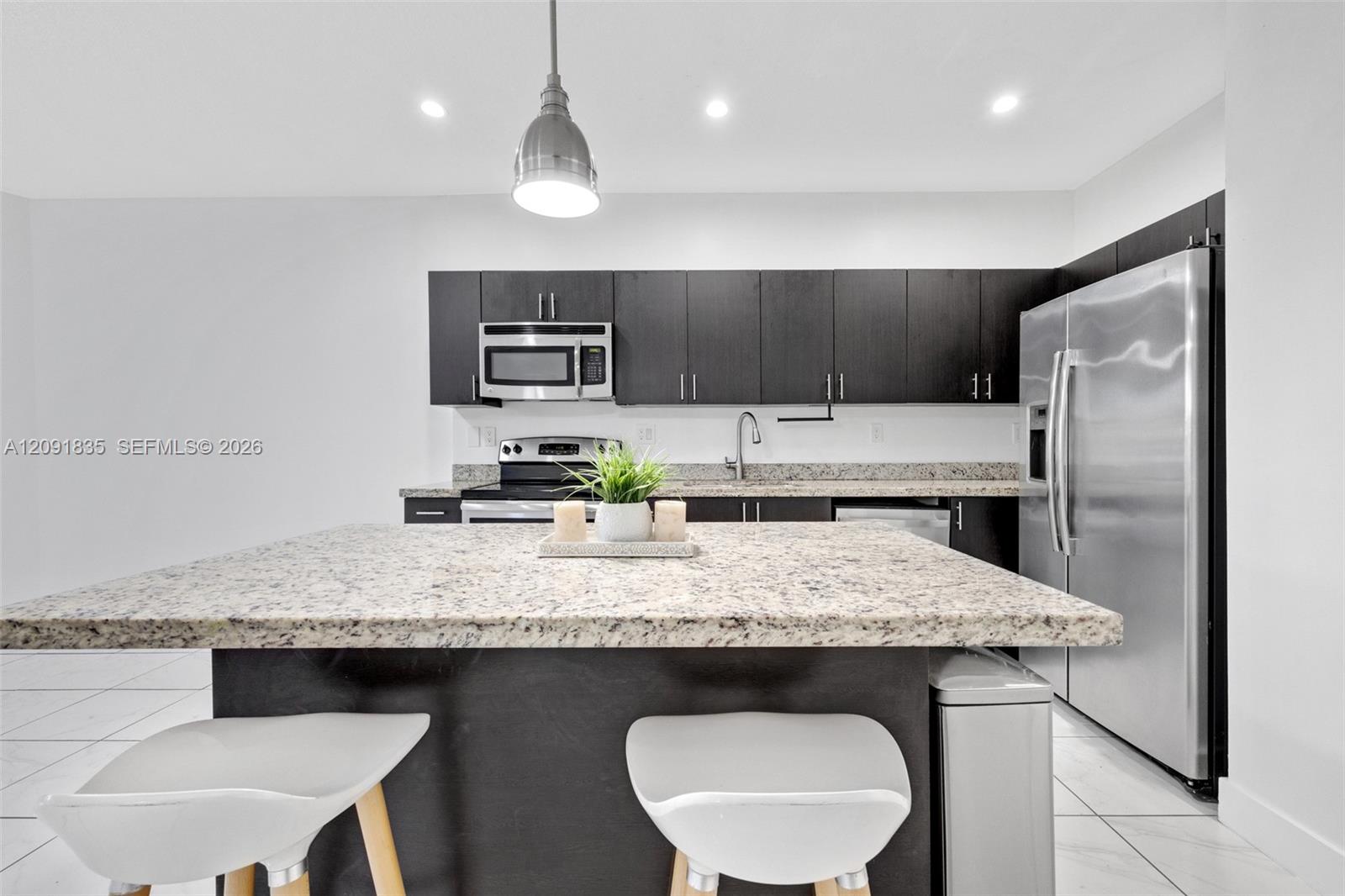 Kitchen featuring dark-toned cabinetry, light-toned stone-finish island countertop, stainless steel appliances, recessed ceiling lighting, and tile flooring
