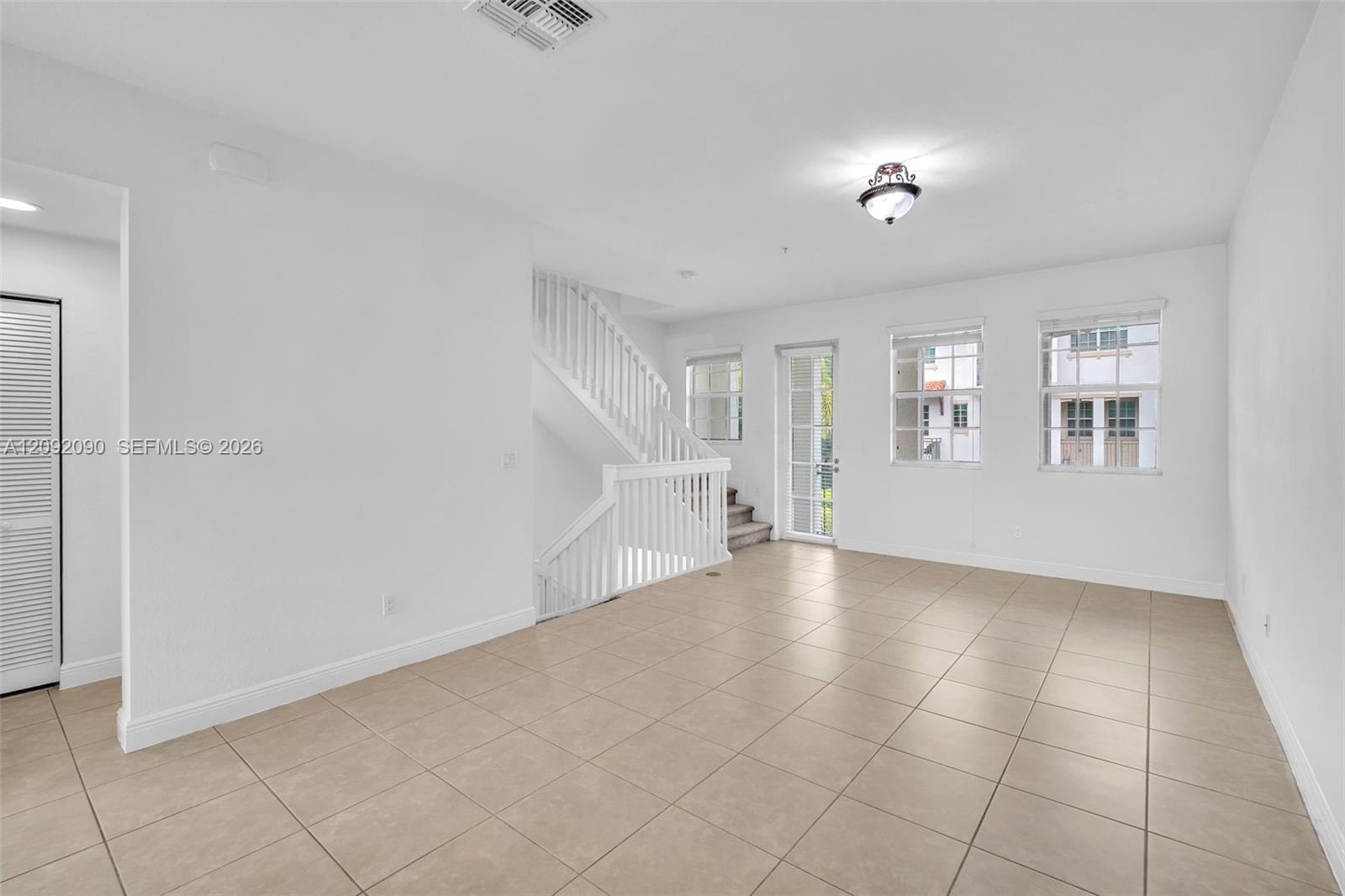 Open-concept interior featuring large-format ceramic tile flooring, a white wooden staircase with balustrades, multiple grid-pane windows, and a glass-paneled exterior door