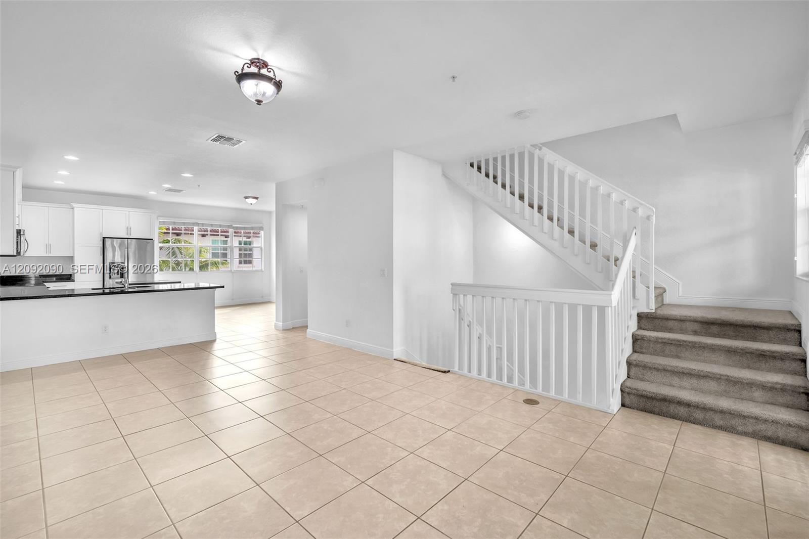 Open-concept interior featuring light-toned tile flooring, white cabinetry, a kitchen island with stone-finish countertops, and a white balustrade staircase with carpeted treads