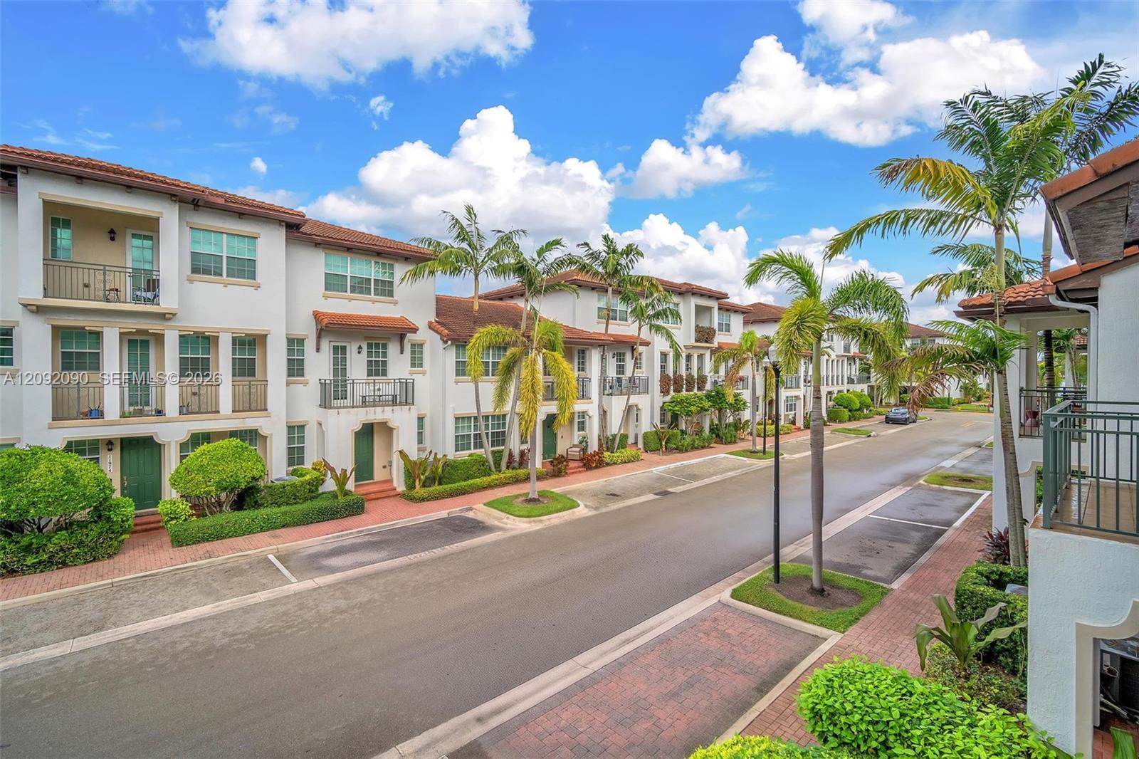 Multi-story stucco townhome exteriors featuring red tile roofing, decorative wrought-iron balconies, private brick-paver walkways, and mature tropical landscaping