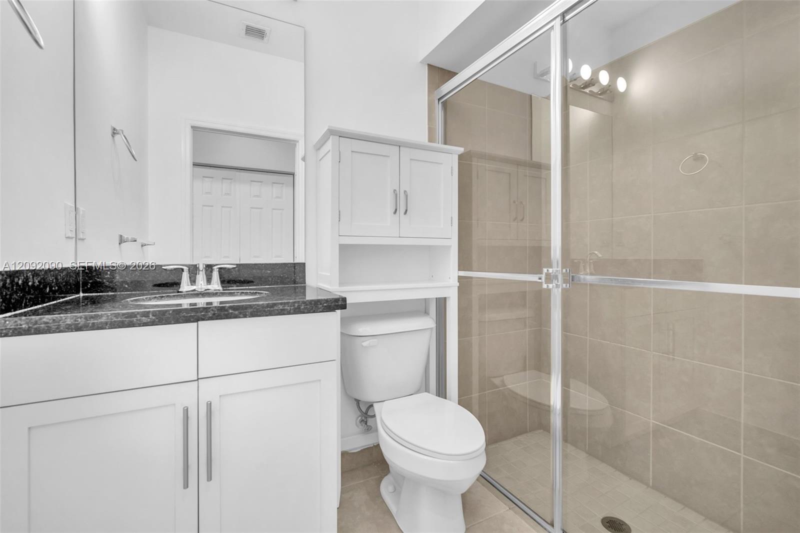 Bathroom featuring a vanity with dark stone countertop, white shaker-style cabinetry, an integrated over-toilet storage unit, and a walk-in shower with tiled surround and glass enclosure