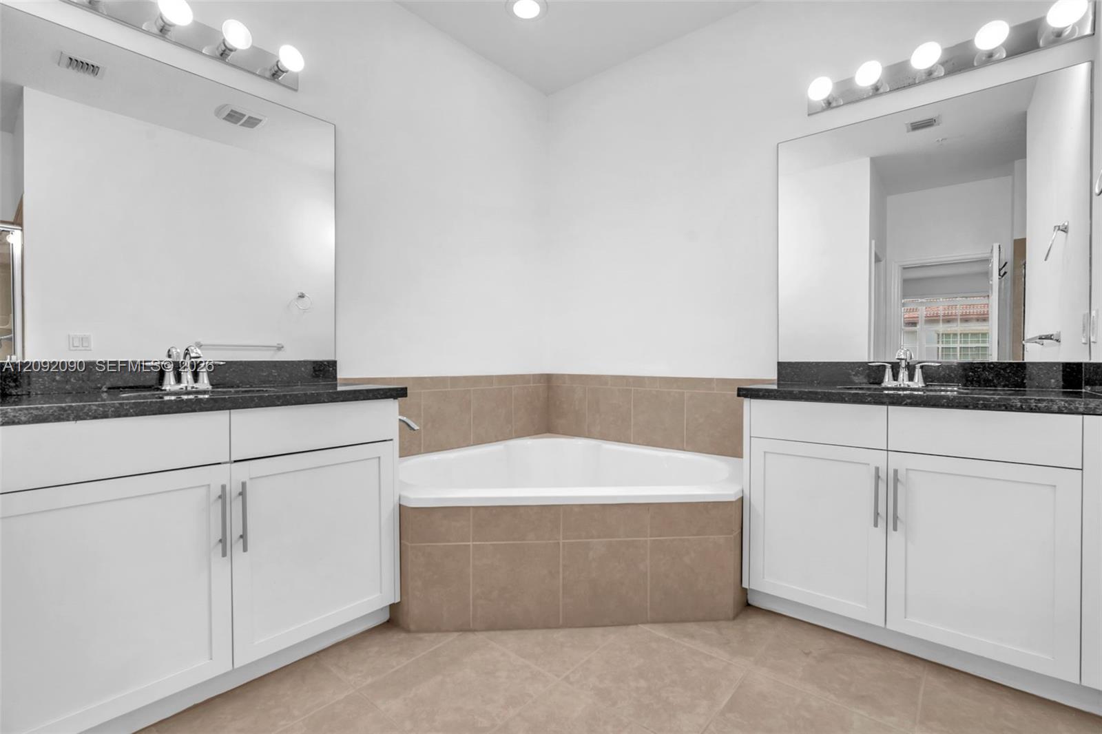Dual white vanities with dark stone countertops, corner soaking tub with tile surround, two large wall-mounted mirrors, and chrome fixtures