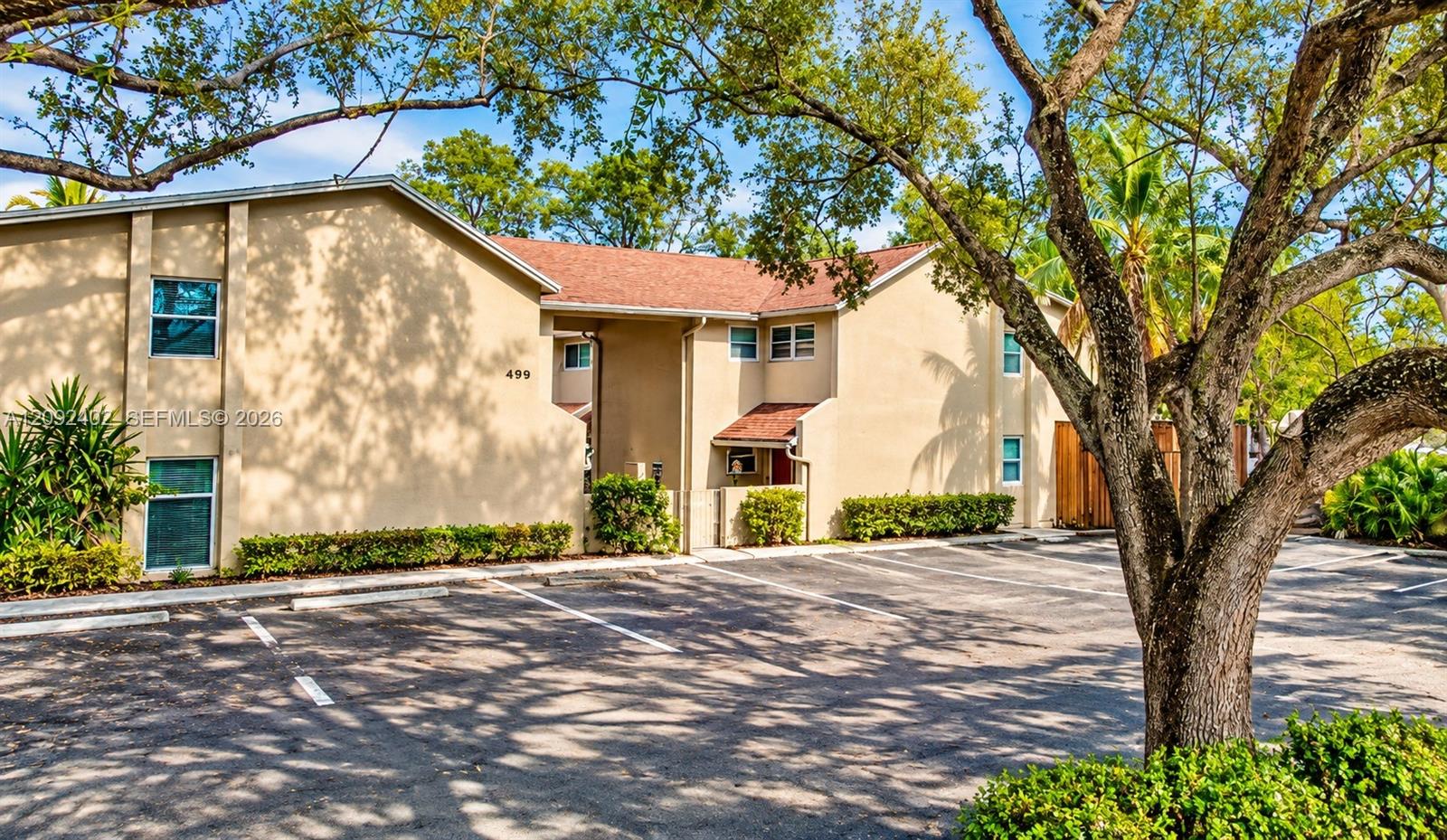 Some landscaping visually staged for illustrative purposes. Exterior building facade featuring a tan stucco finish, multi-pitched shingled roof, asphalt-paved parking area.