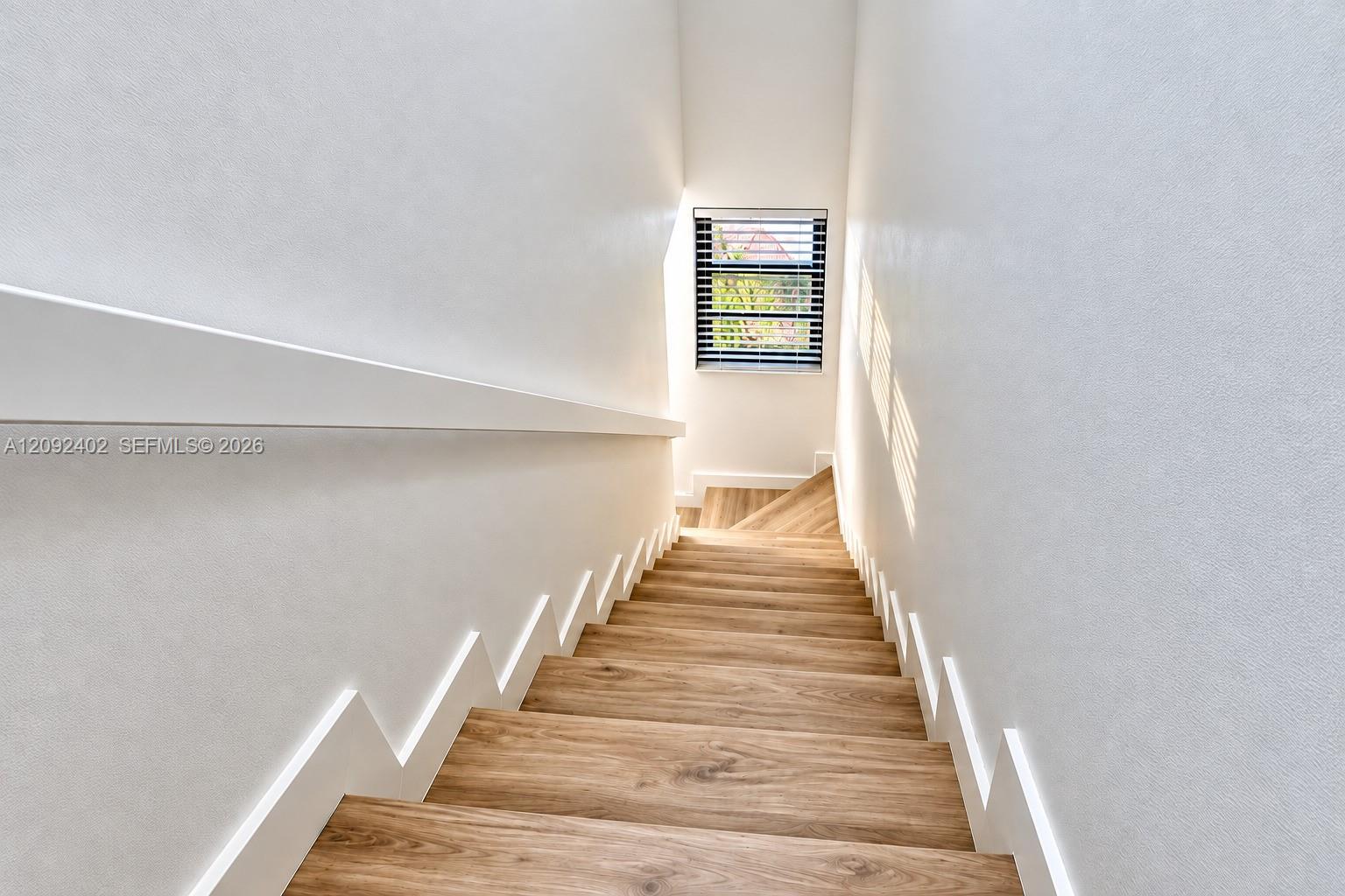 Staircase featuring wood-finish treads, white stringer detailing, a wall-mounted handrail, and a window with horizontal blinds
