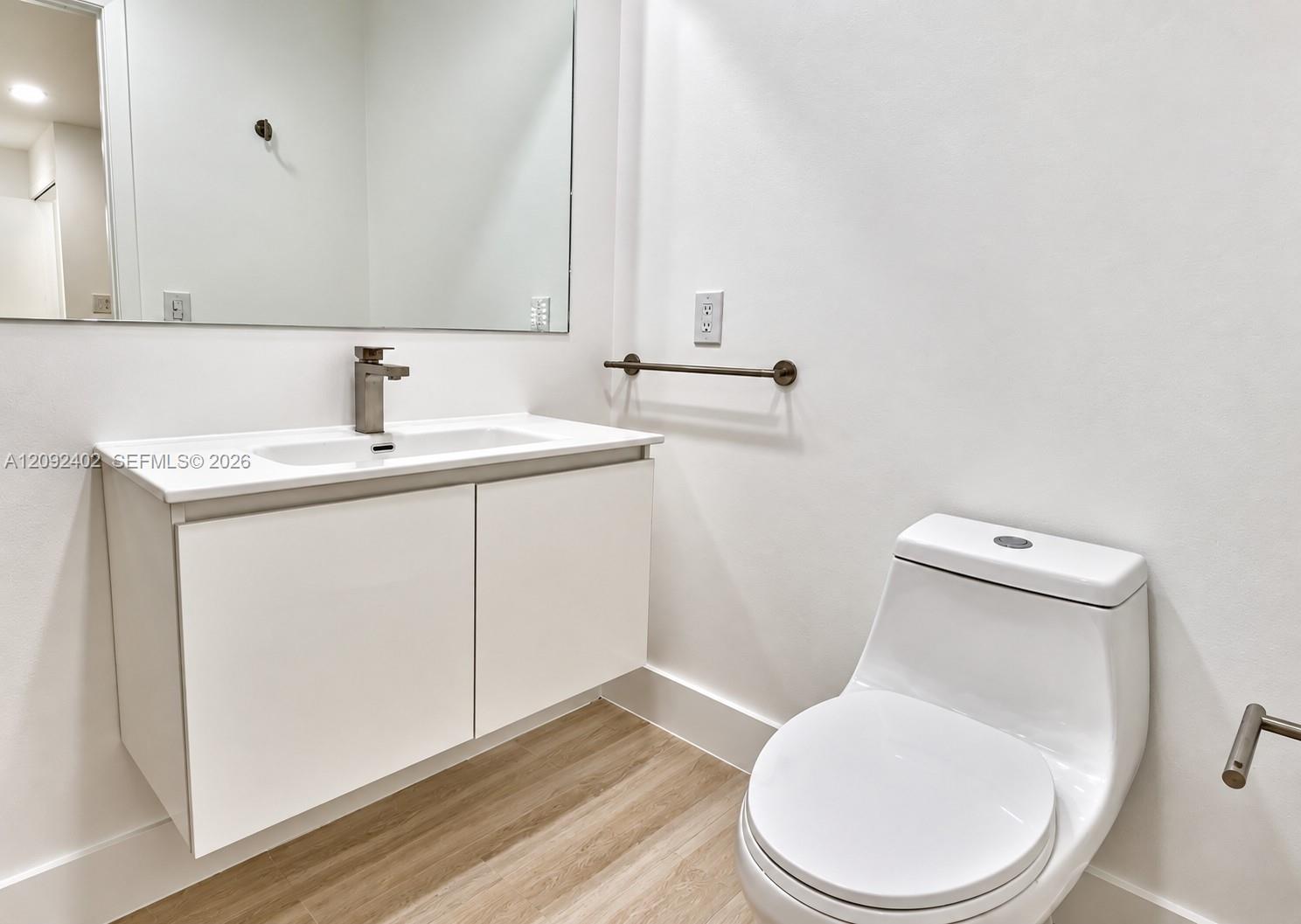 Floating vanity with a white countertop and integrated sink, wall-mounted faucet, expansive frameless mirror, one-piece dual-flush toilet, and wood-finish flooring
