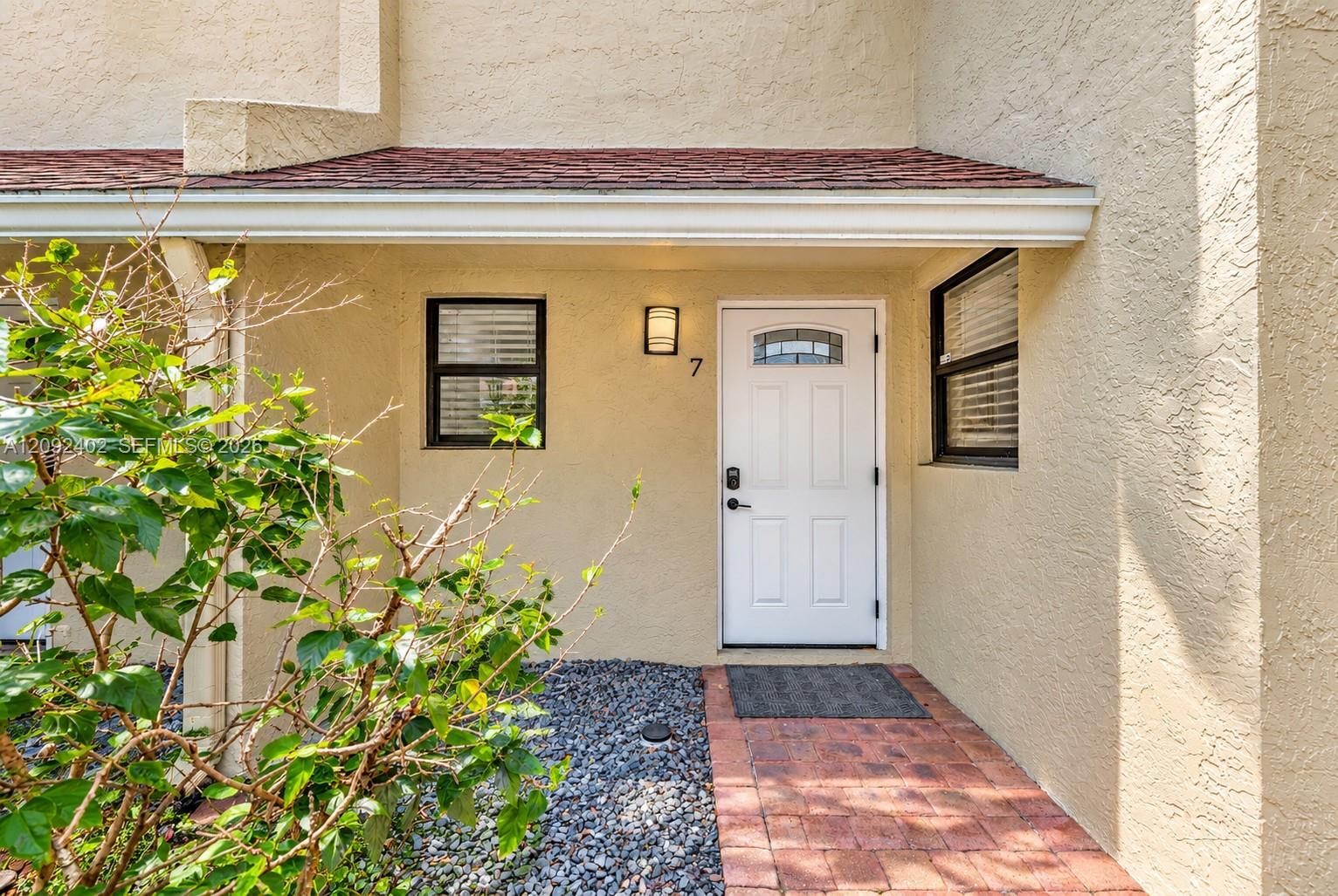 Stucco exterior entrance featuring a brick-paver walkway, red shingle roof overhang, white door with a glass inlay, and wall-mounted exterior lighting