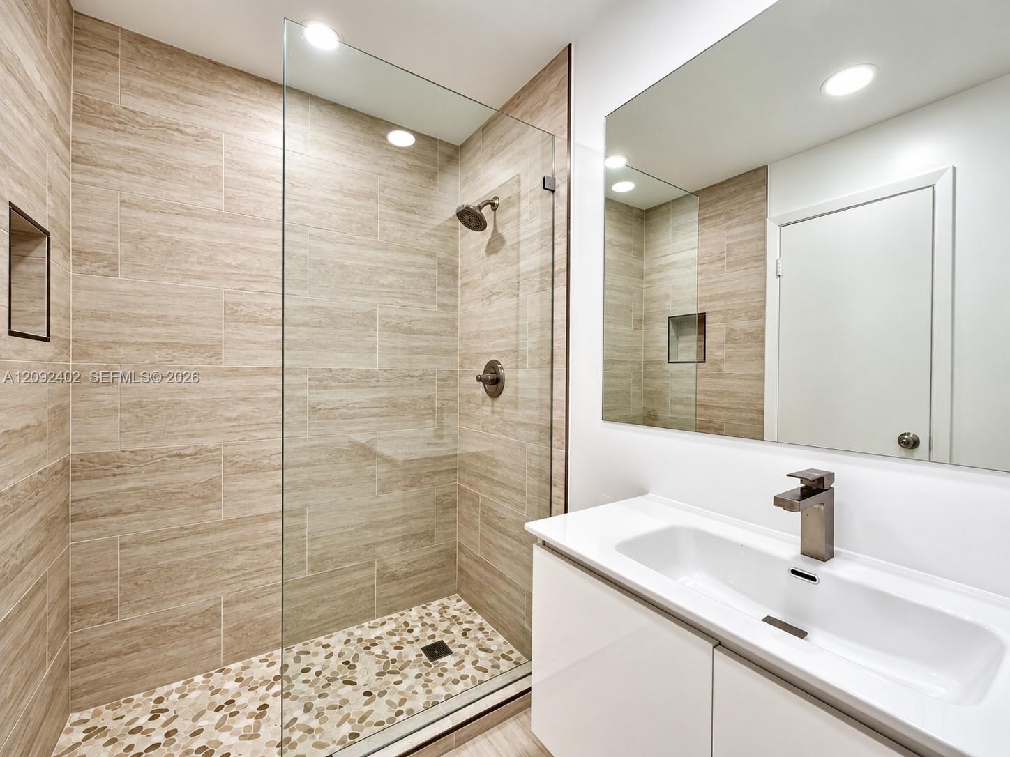 Walk-in shower featuring floor-to-ceiling textured tile, a river-stone floor, and a frameless glass enclosure, paired with a white vanity featuring a rectangular basin, stone countertop, and brushed metal fixtures