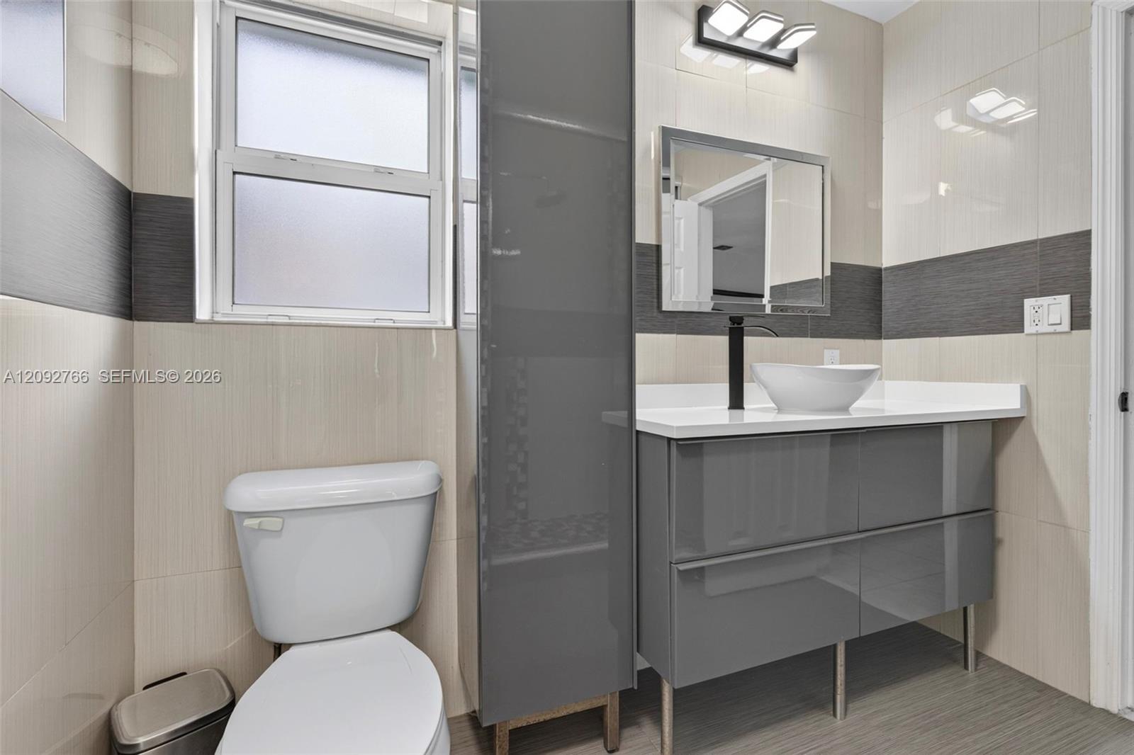 Modern floating vanity featuring a glossy grey finish, white countertop, vessel sink, and matte black faucet, complemented by full-height wall tiling with contrasting horizontal banding