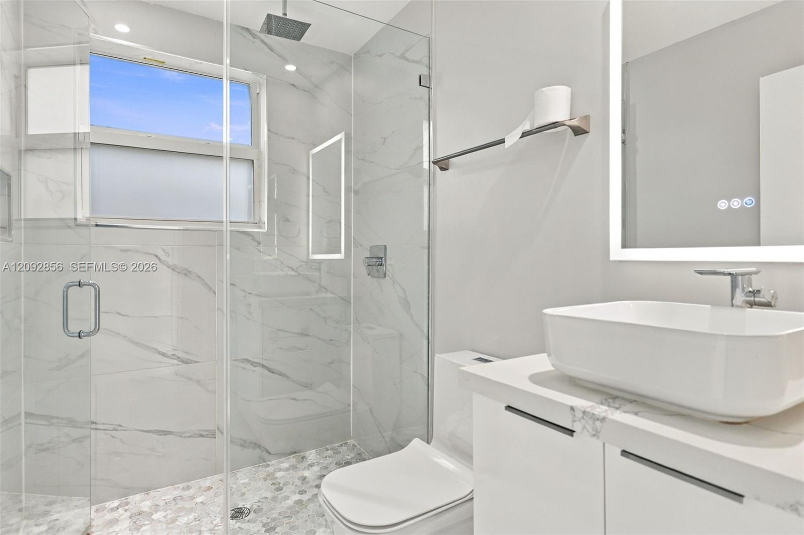 2nd bathroom first floor - Cabana Bathroom featuring a glass-enclosed shower with floor-to-ceiling stone-patterned tile, a rainfall showerhead, a pebble-finish shower floor, a floating vanity with an integrated vessel sink, and an illuminated mirror