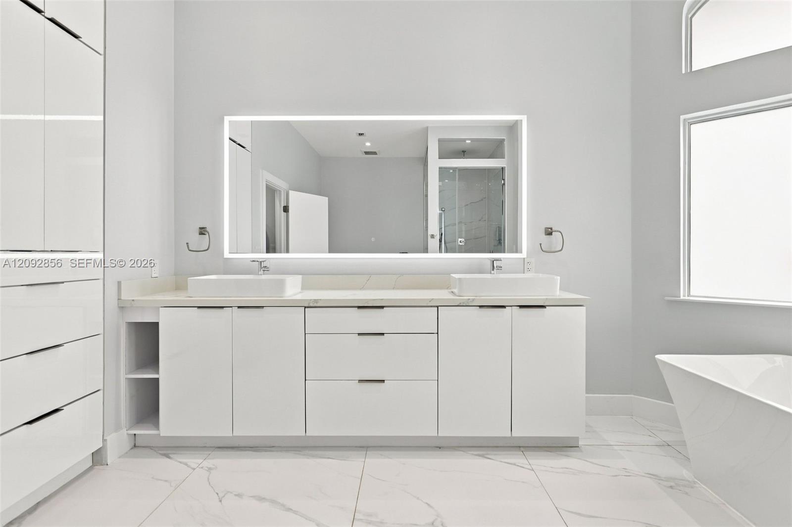 Bathroom featuring a dual-sink vanity with matte white cabinetry, stone-surface countertop, rectangular vessel sinks, an expansive LED-backlit mirror, and large-format porcelain floor tile
