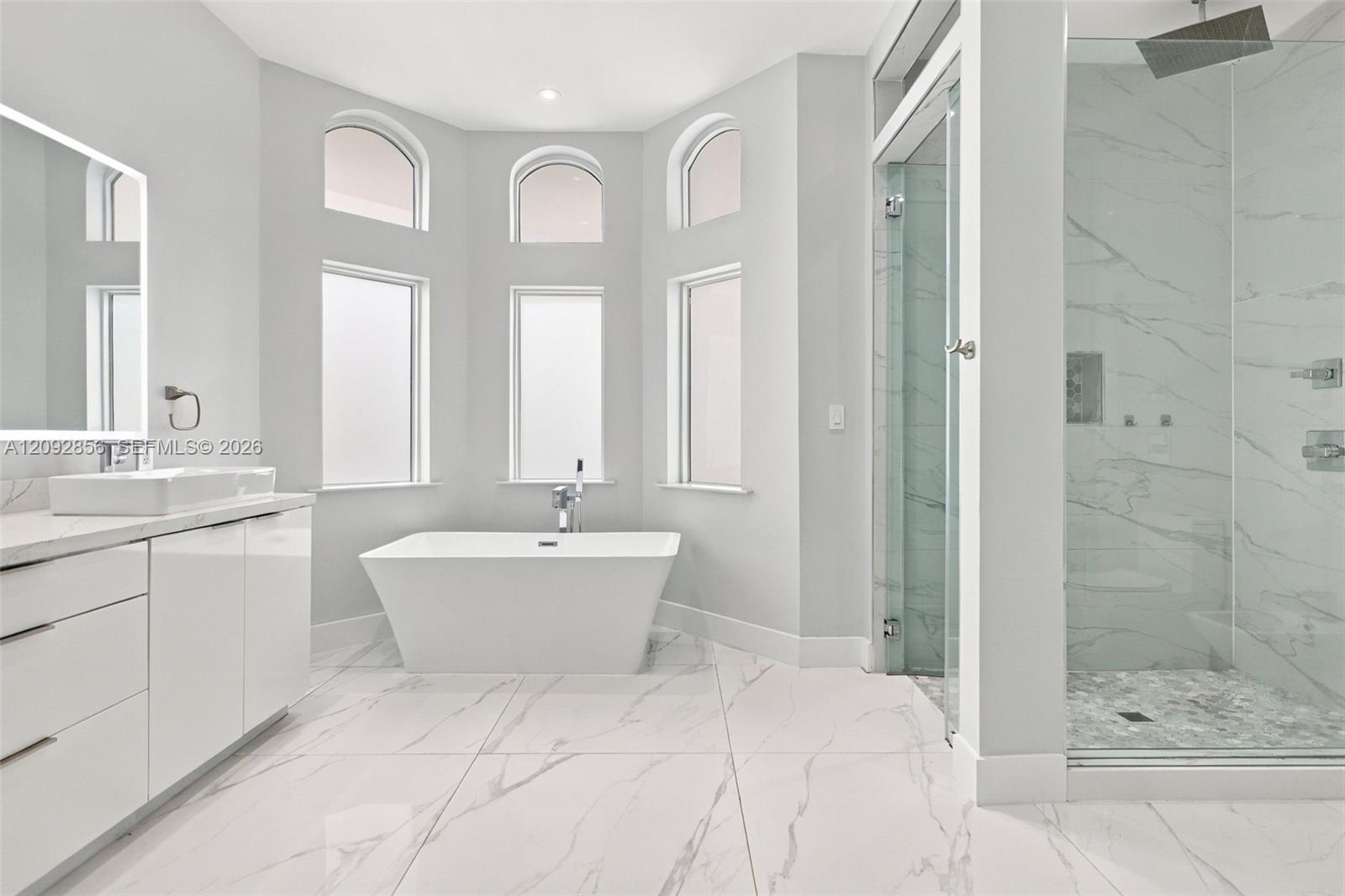 Freestanding soaking tub set in a bay alcove with arched windows, large-format tile flooring, frameless glass-enclosed shower, and sleek vanity with a vessel sink