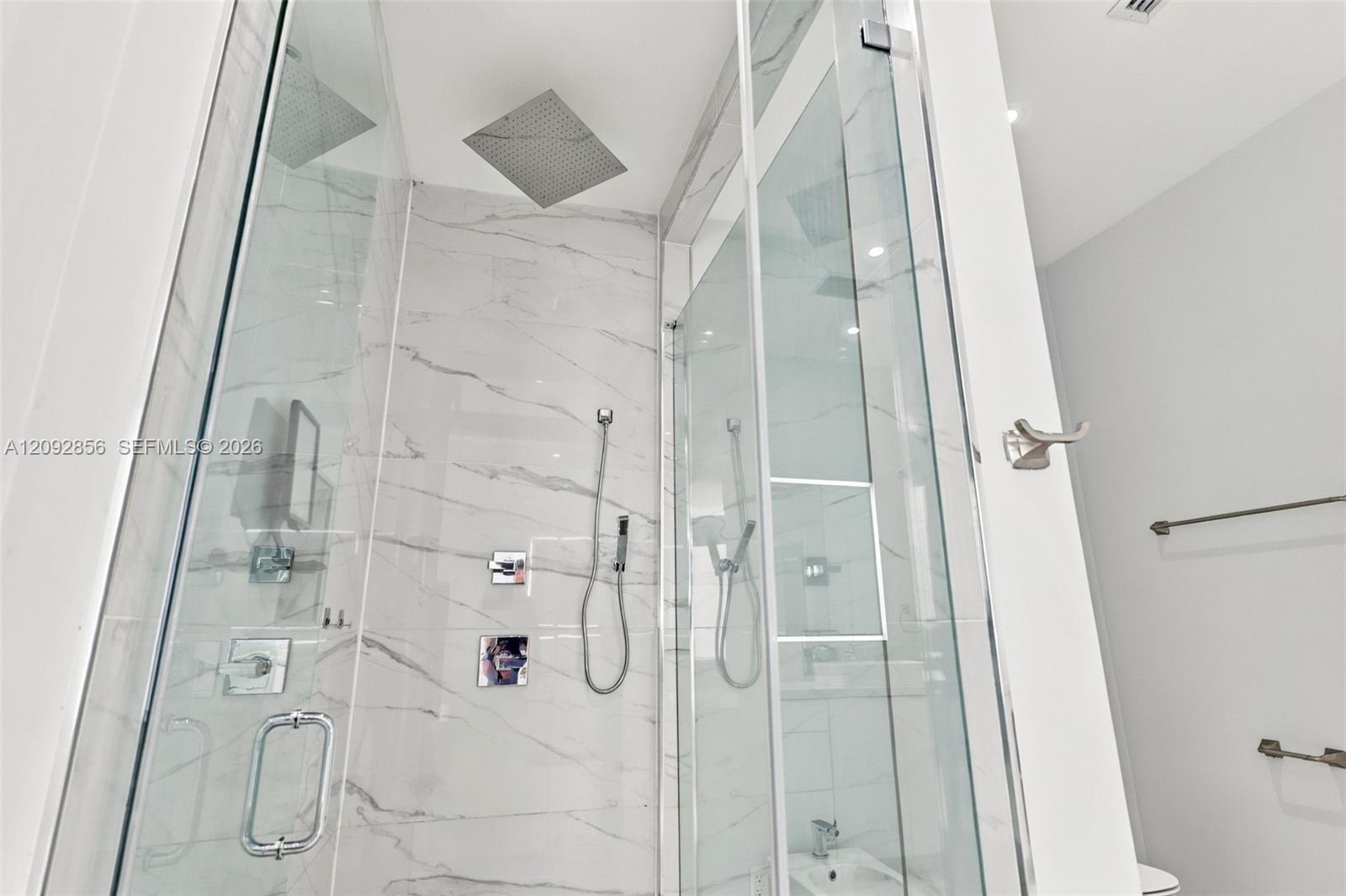 Glass-enclosed walk-in shower featuring floor-to-ceiling stone-finish tile, a square rainfall showerhead, a separate handheld sprayer, and polished chrome fixtures