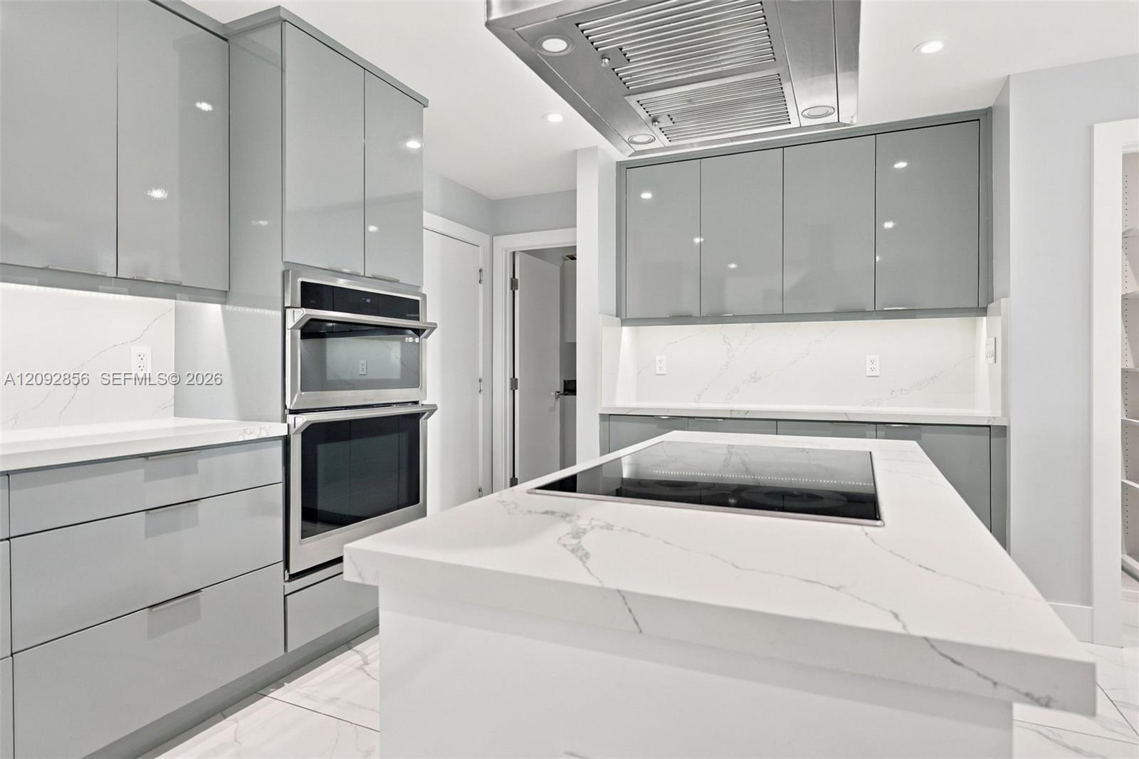 Modern kitchen featuring high-gloss grey cabinetry, stone-surface countertops with waterfall edge, integrated induction cooktop, stainless steel built-in double oven, and industrial-style range hood