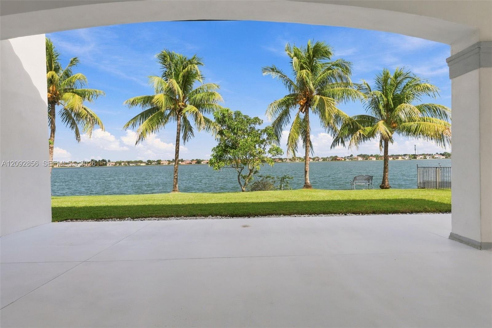 Covered patio featuring a concrete floor, arched structural support, manicured lawn, palm trees, and water frontage