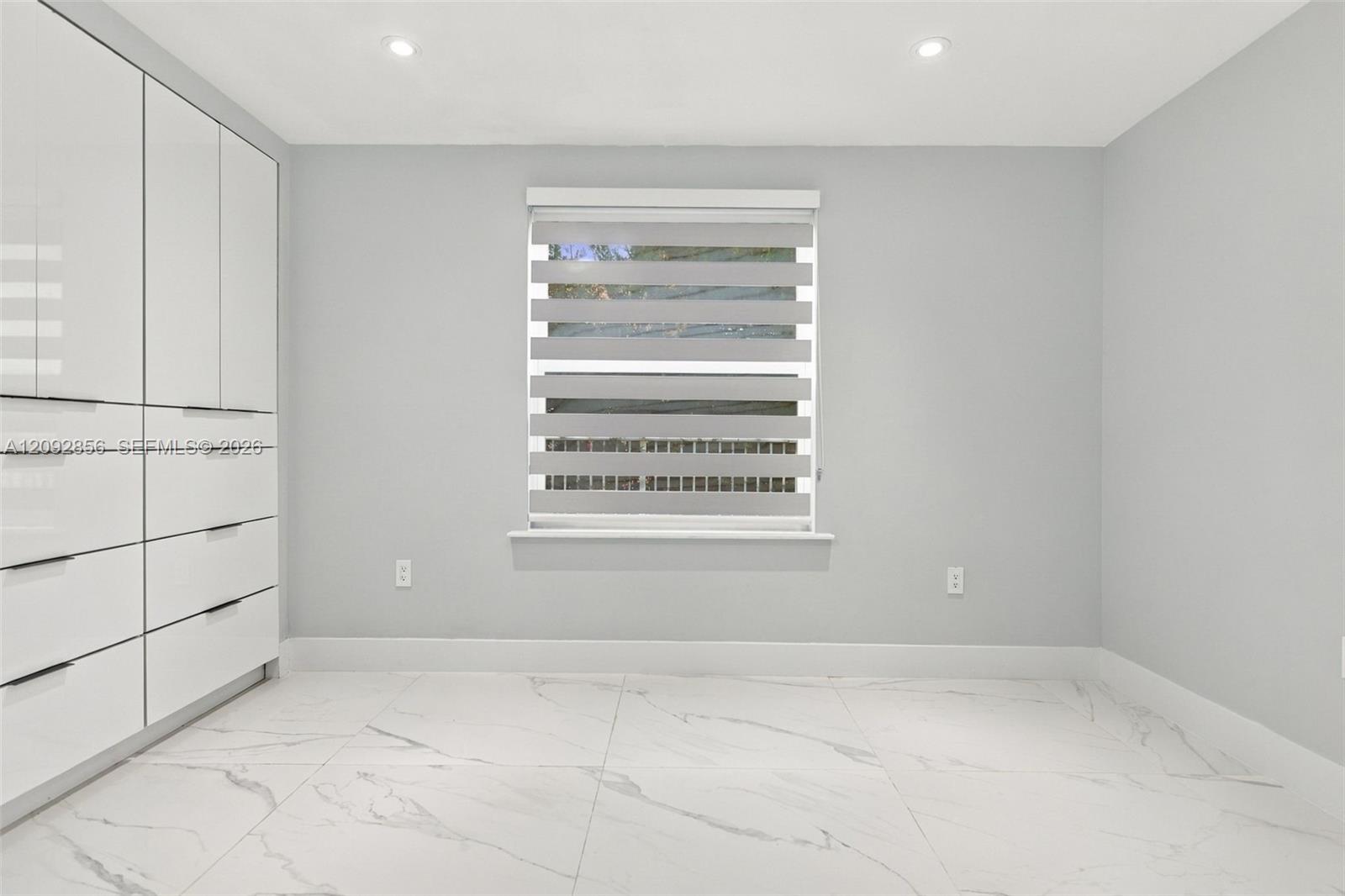 2nd bedroom - 1st floor - Built-in floor-to-ceiling cabinetry with white high-gloss finish, large-format tile flooring with veining detail, recessed lighting, and a window featuring a dual-layer roller shade