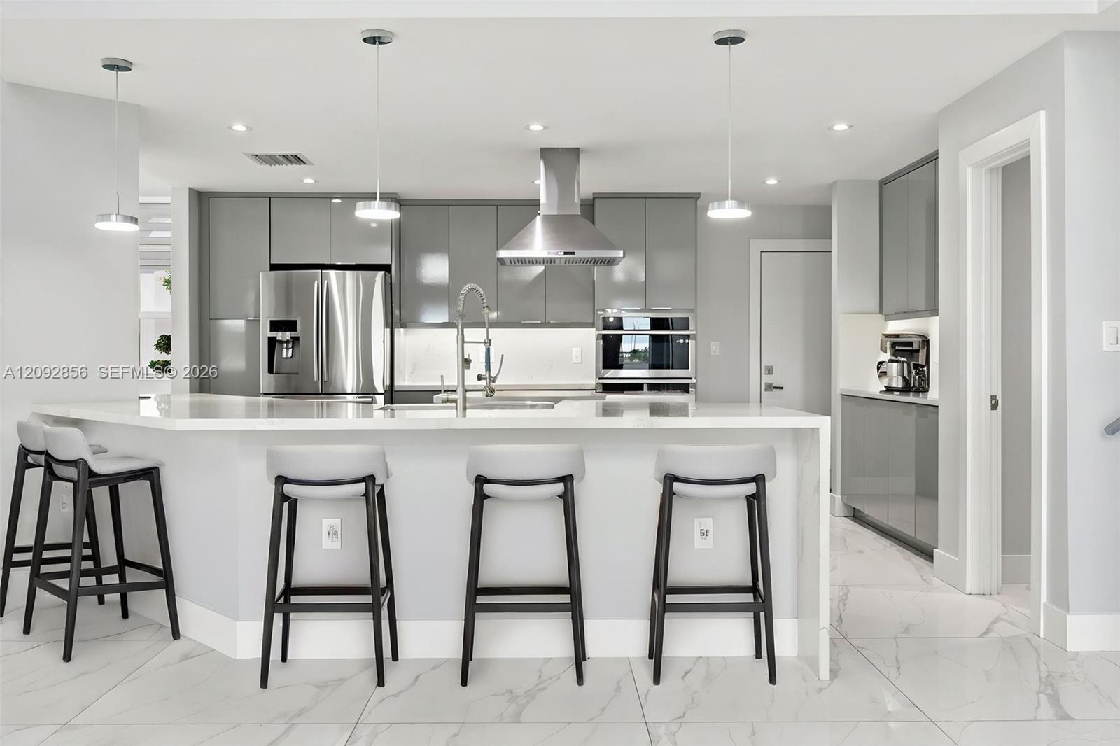Virtually staged - itchen featuring glossy grey cabinetry, stainless steel appliances, stone-surface waterfall island with a gooseneck faucet, pendant lighting, and large-format tile flooring