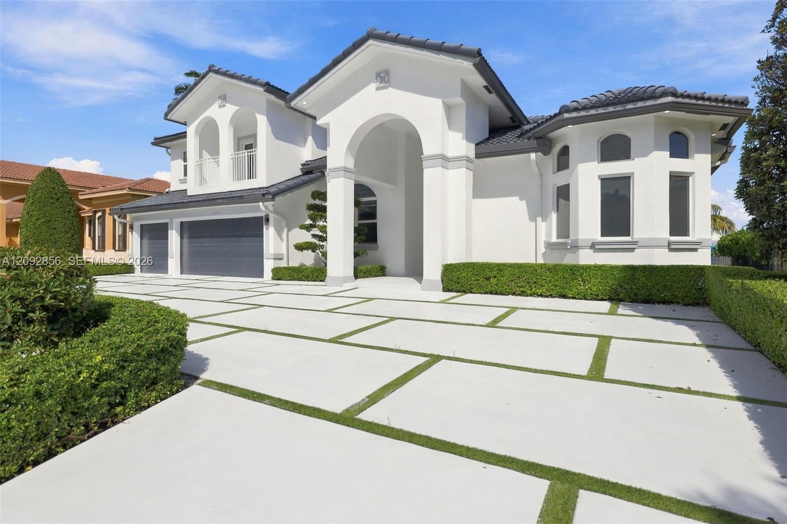 Stucco exterior with a grand arched entryway, multi-car garage, tiled roof, and paver driveway with turf accents